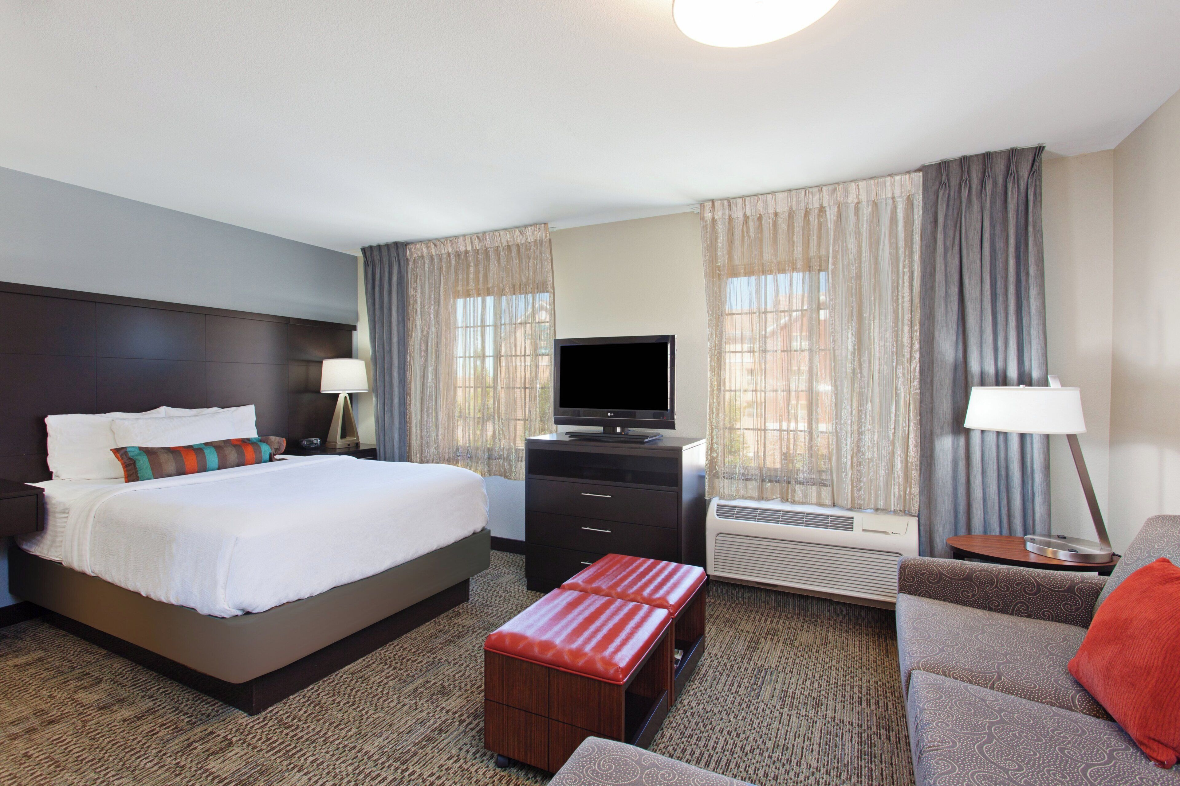 A hotel room with a bed, a TV, a desk, a chair, a sofa, and a window with curtains.