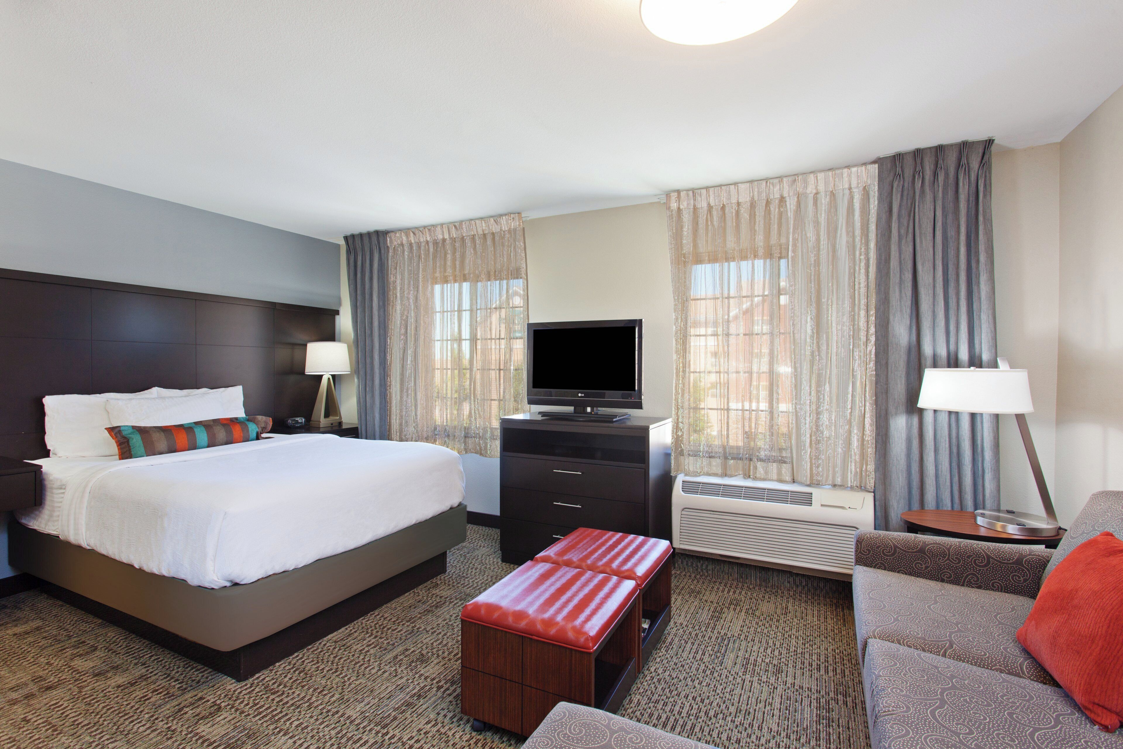 A hotel room with a bed, a TV, a desk, a chair, a sofa, and an air conditioning unit.