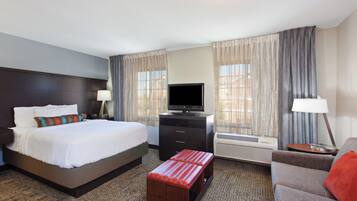 A hotel room with a bed, a TV, a desk, a chair, a sofa, and an air conditioning unit.