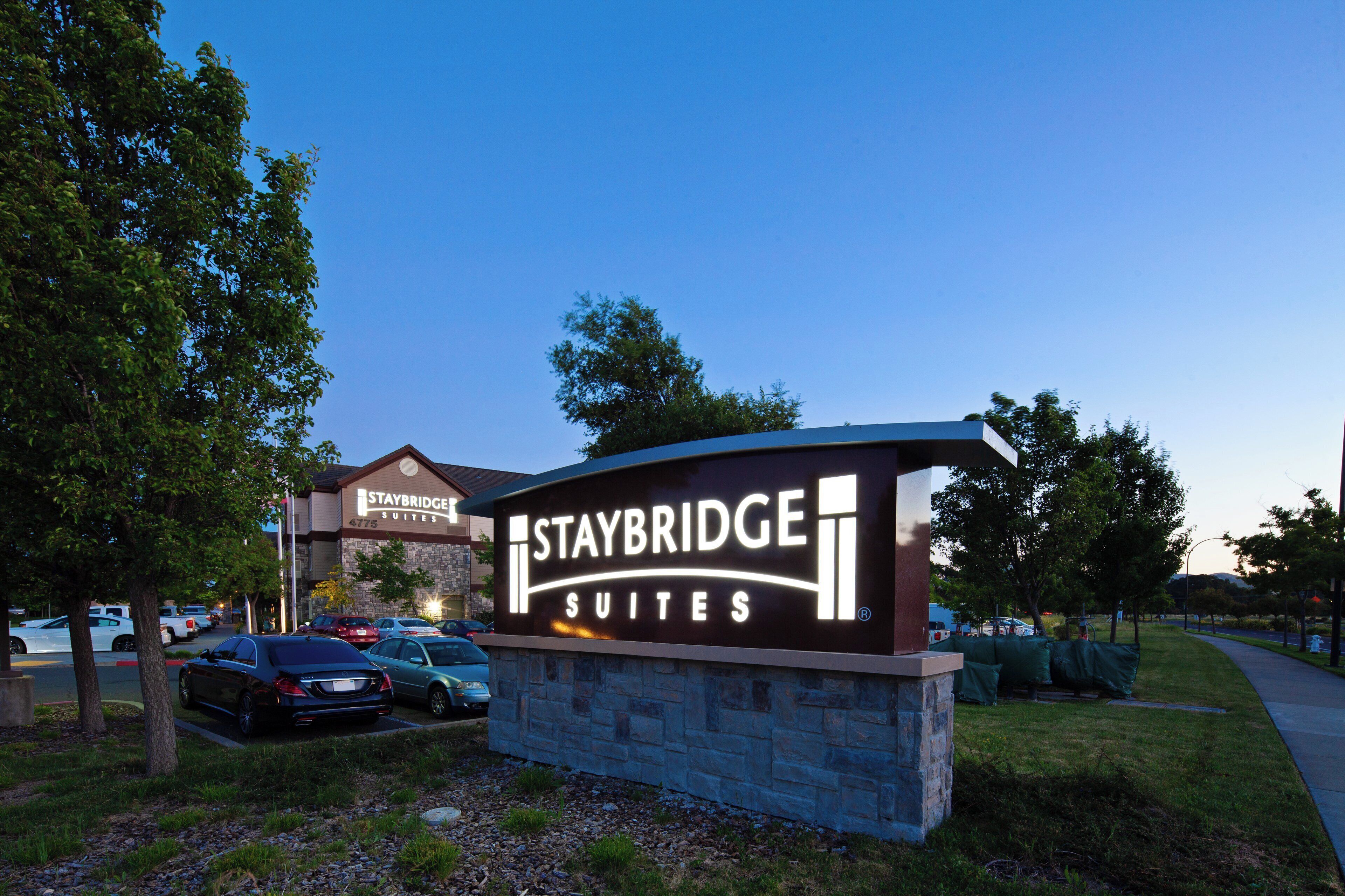 A Staybridge Suites sign with a stone base, surrounded by parked cars and trees.