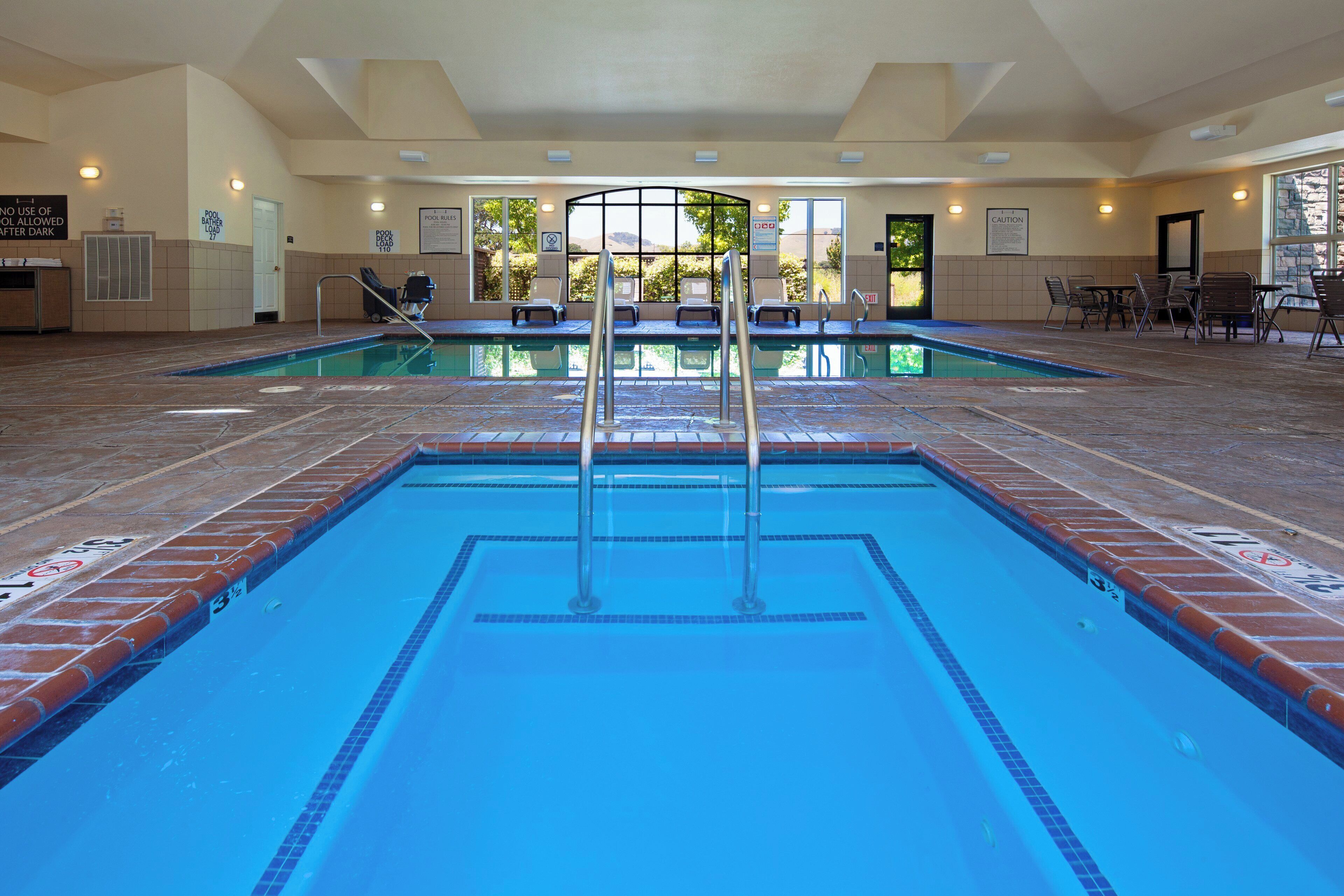 Una gran piscina interior con agua cristalina y azul, rodeada de sillones de descanso y una zona cubierta con asientos.