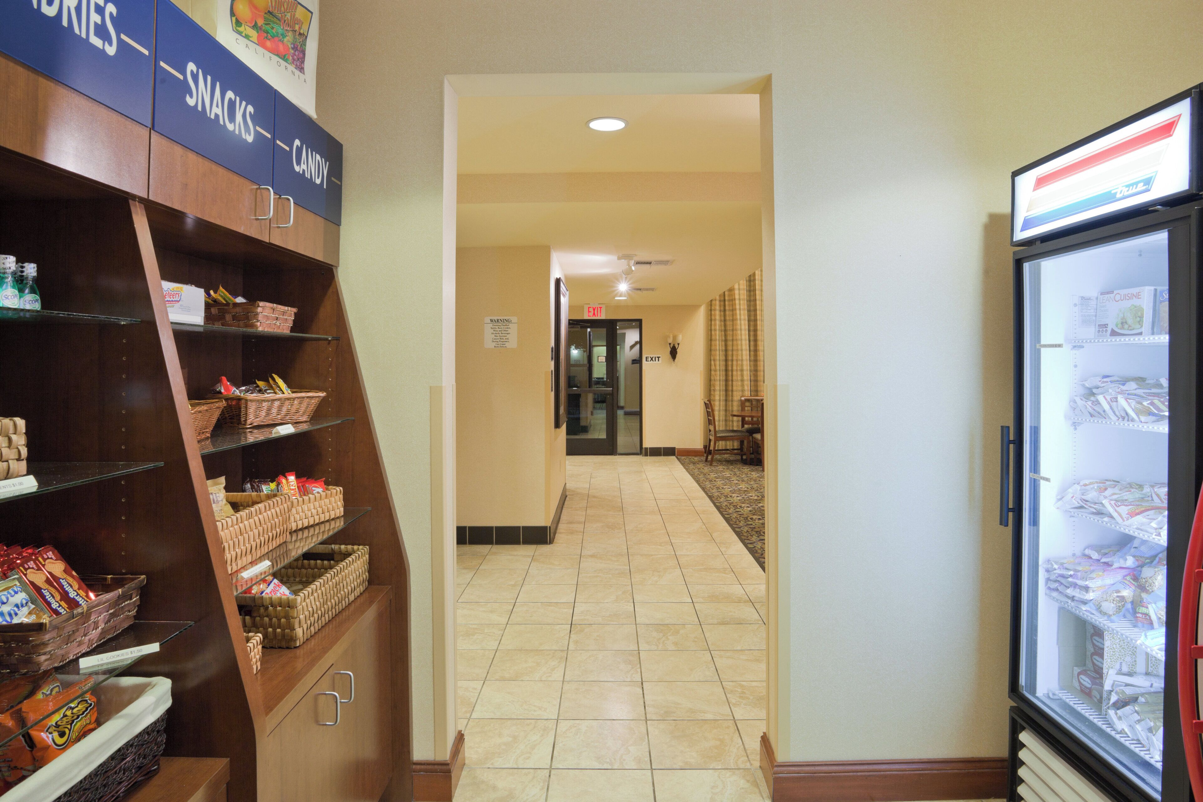 A hotel corridor with snack and candy shelves, a refrigerator stocked with beverages, and an exit sign.