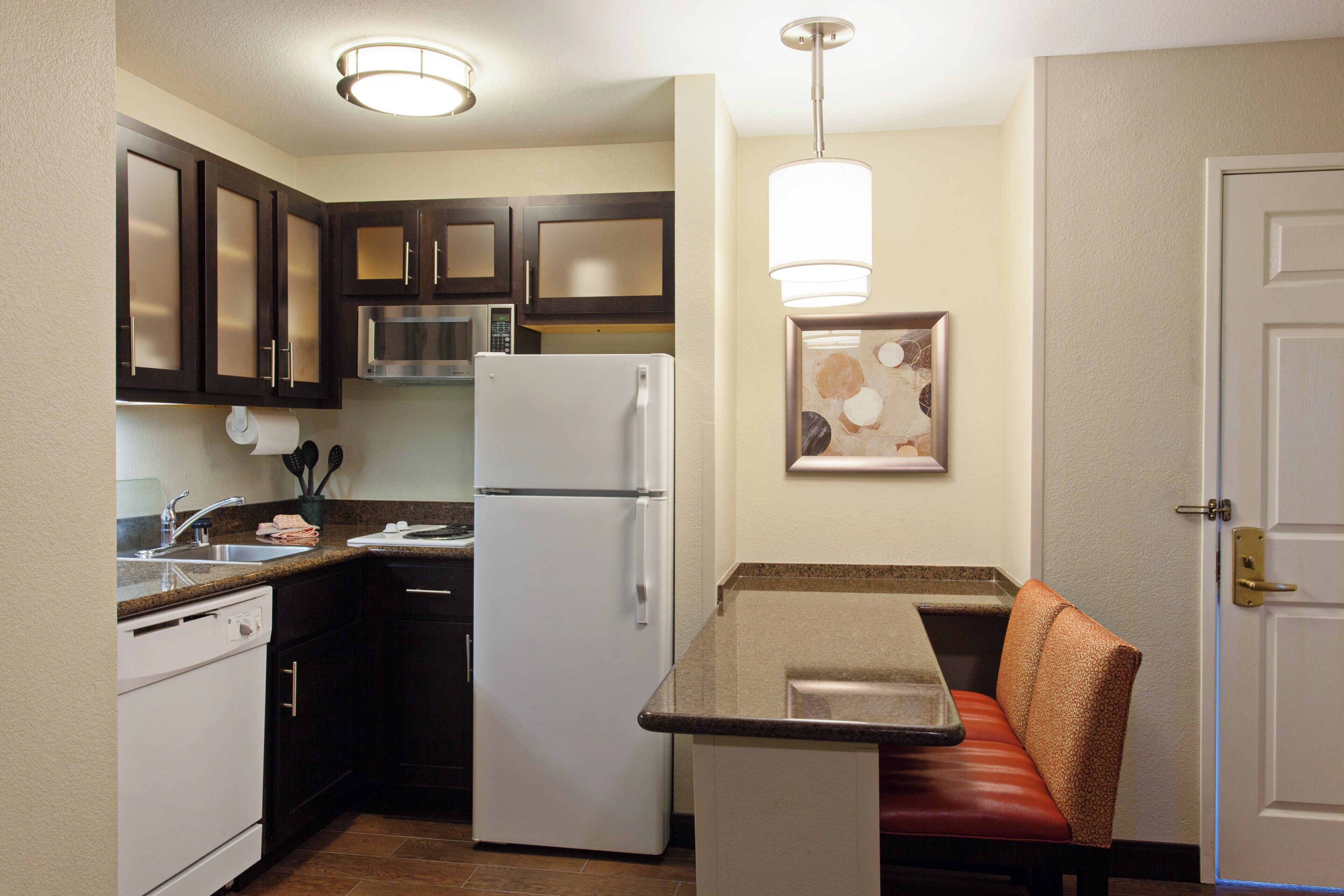 A compact kitchen with dark cabinets, a white refrigerator, a microwave, and a sink. There's a small seating area with a cushion and a framed picture on the wall.
