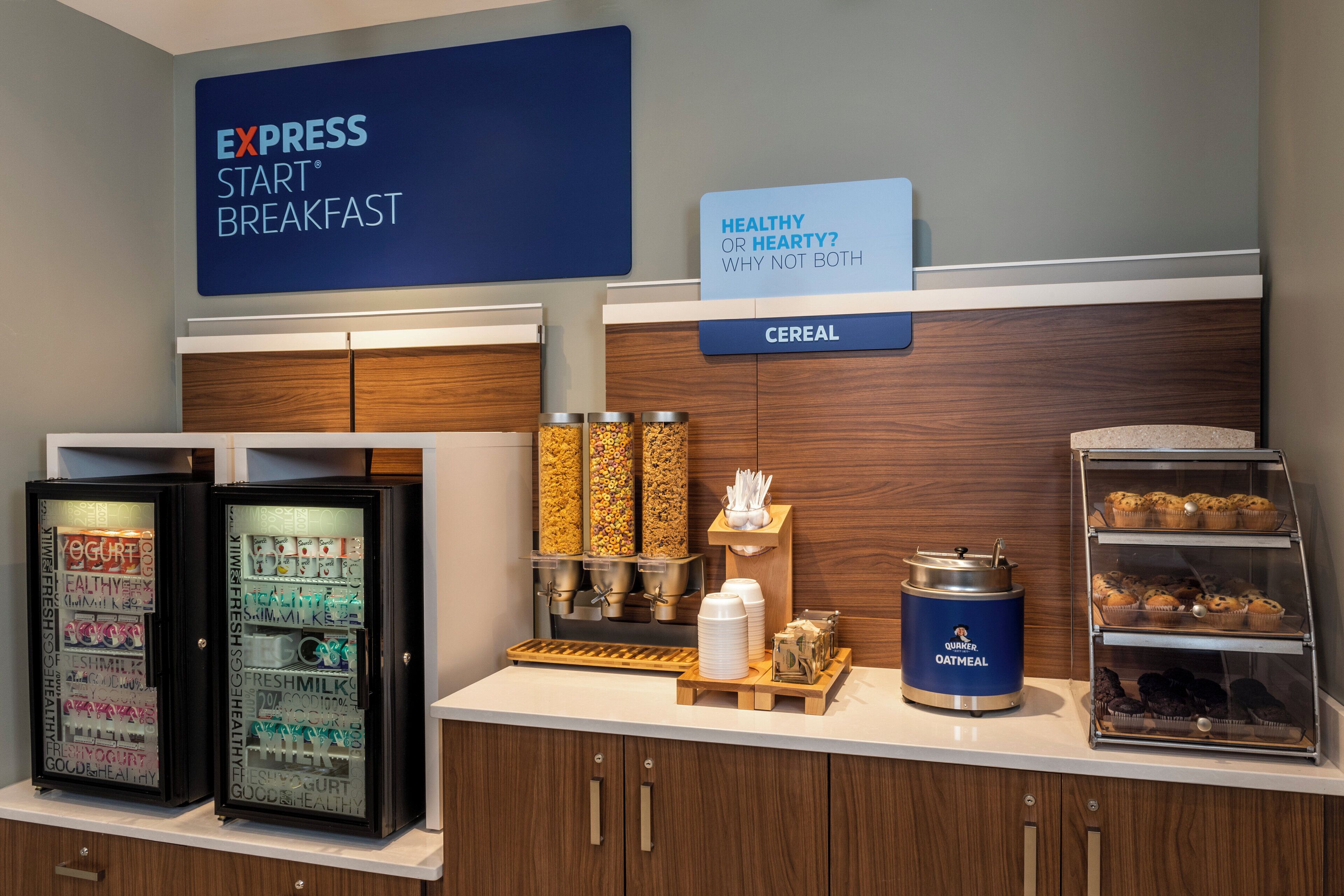 Estación de desayuno con un letrero que dice 'EXPRESS START BREAKFAST' y un letrero que pregunta '¿SALUDABLE O SUSTANCIOSO? ¿POR QUÉ NO AMBOS'. Hay dispensadores de cereal, un refrigerador de yogurt y un dispensador de Quaker Oatmeal.