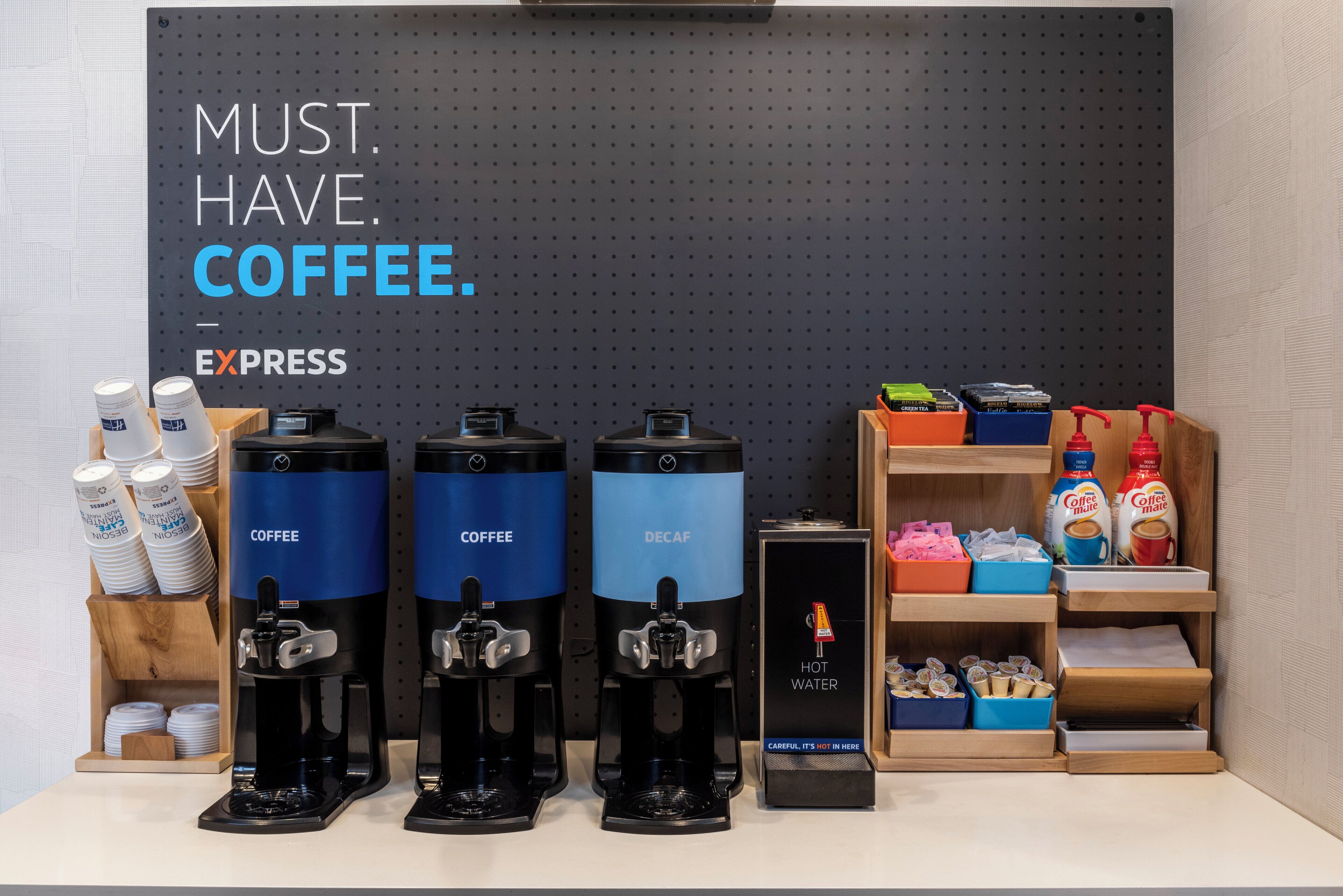 Una estación de café con tres dispensadores de café, un dispensador de agua y una estantería con productos de café.