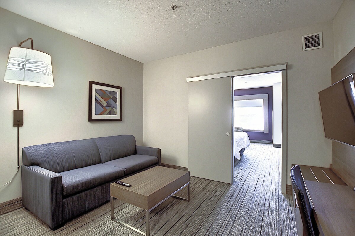 Habitación de hotel con sofá, una mesita, lámpara, un cuadro enmarcado y una puerta que da a otra habitación.