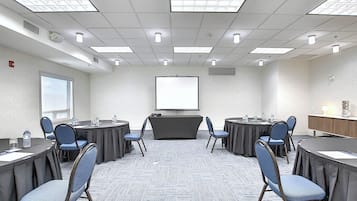 Un salón de conferencias con una pantalla grande, varias mesas con sillas y piso alfombrado.