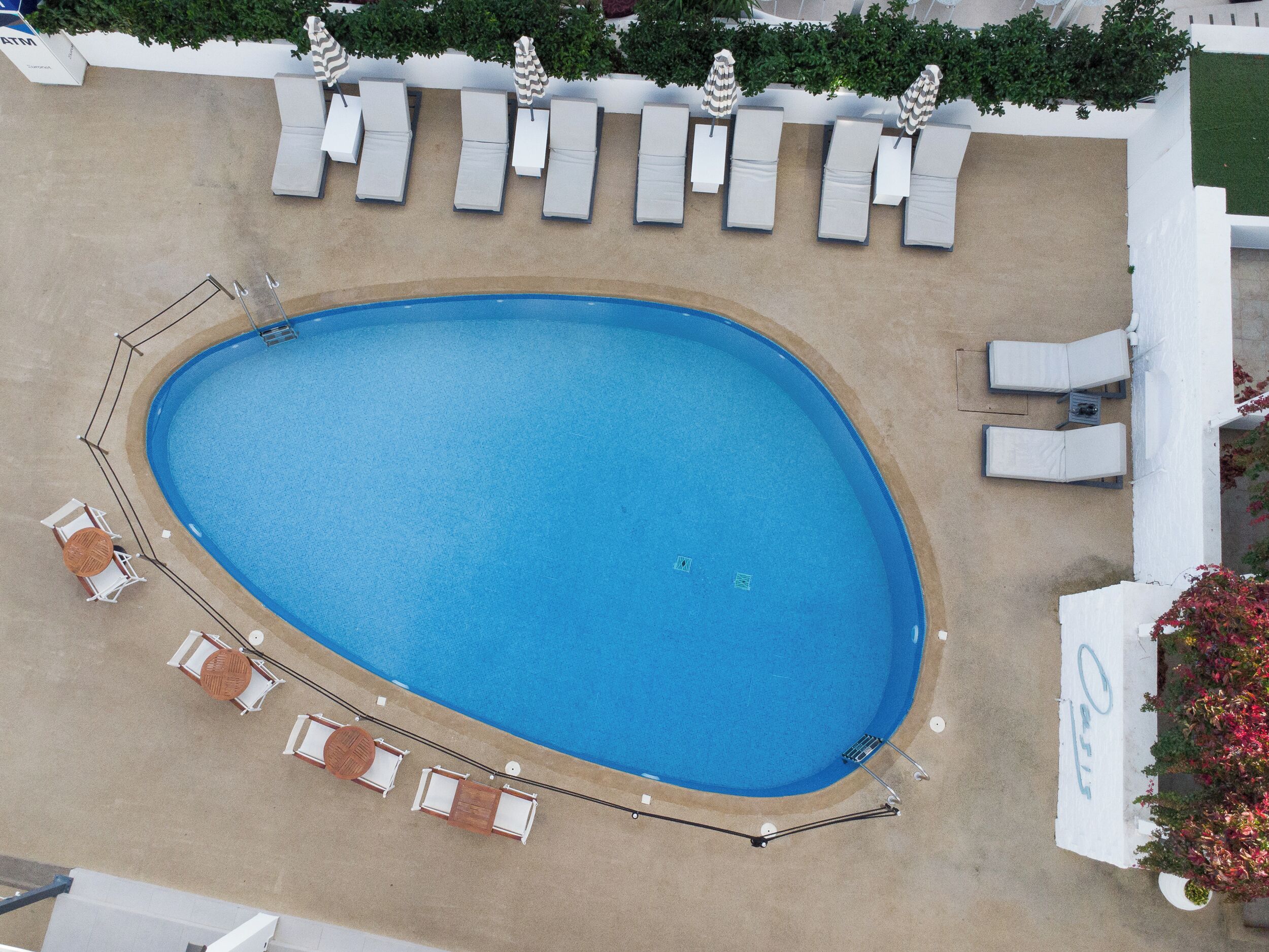 An aerial view of a swimming pool with sun loungers and umbrellas, surrounded by a white wall and greenery.