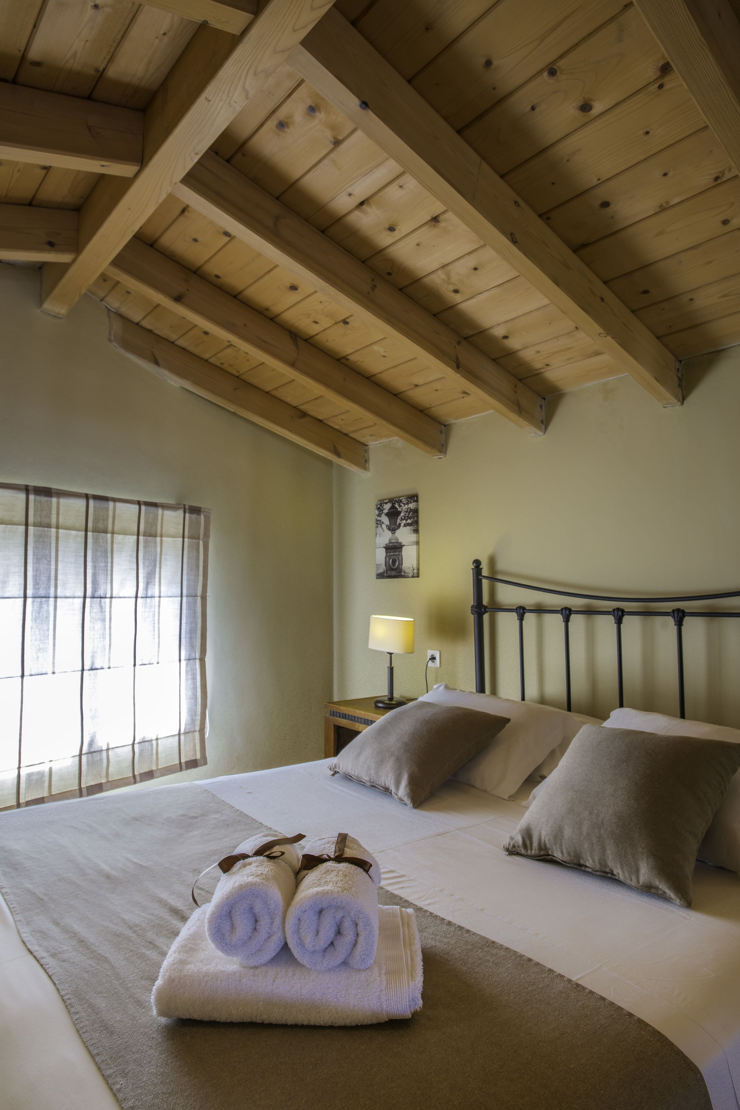 Una cama bien hecha con toallas enrolladas, techo de madera, ventana con persianas y una lámpara de noche.