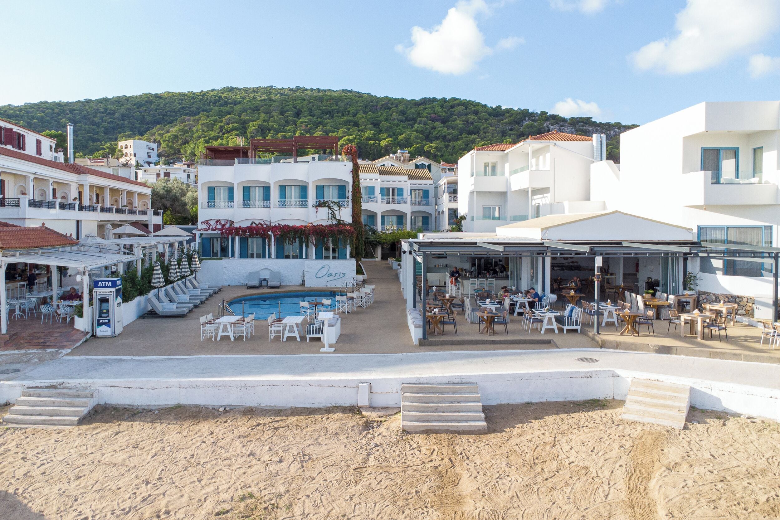 A beachfront hotel with a pool, outdoor seating, and a view of the hillside.