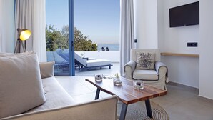 A modern living room with a sofa, a coffee table, a chair, and a TV mounted on the wall. There is a large window with a view of the sea and a balcony with a sun lounger. - Oasis Beach Hotel (Agistri)