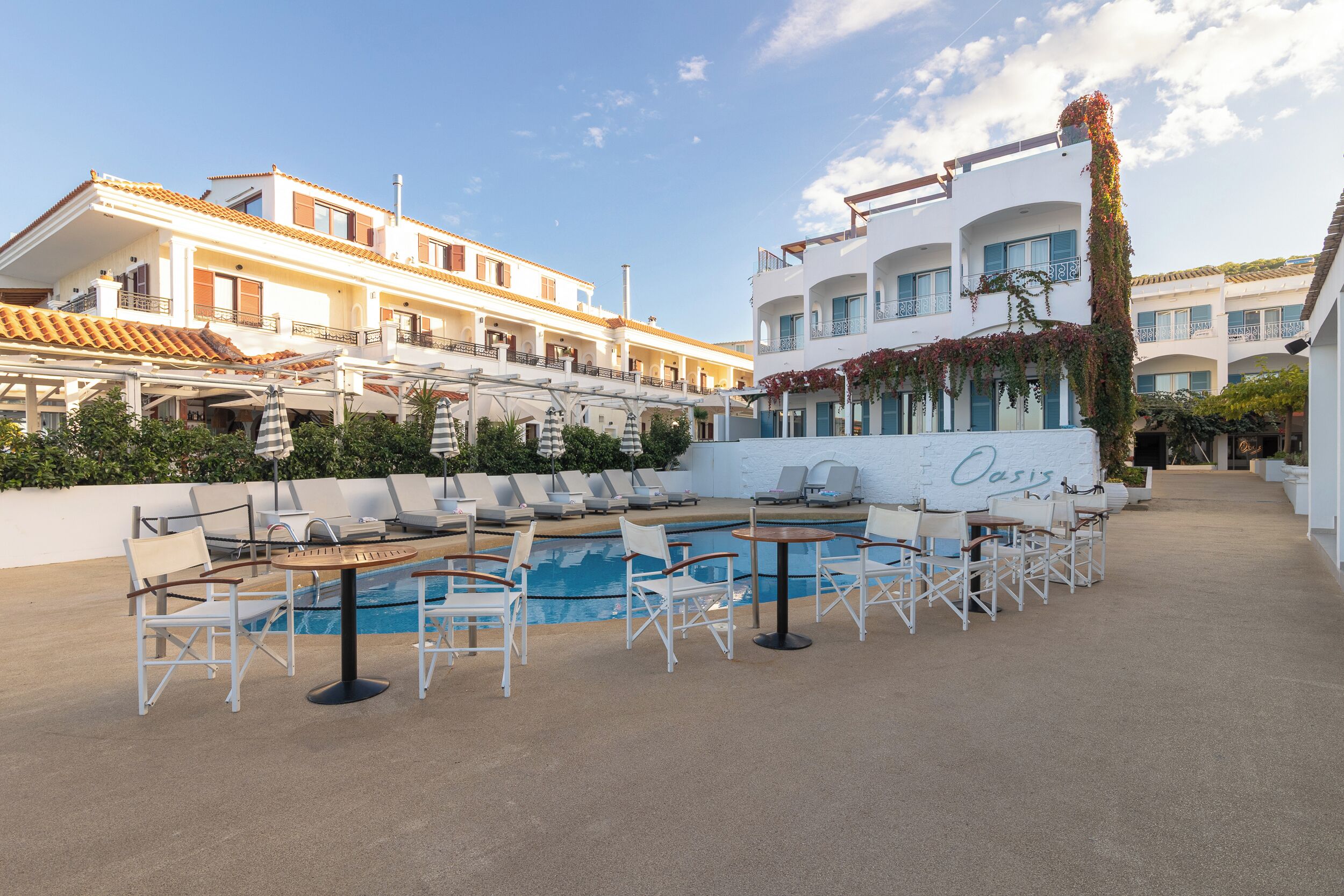A rooftop pool area with lounge chairs and tables, surrounded by buildings with balconies and a clear sky.