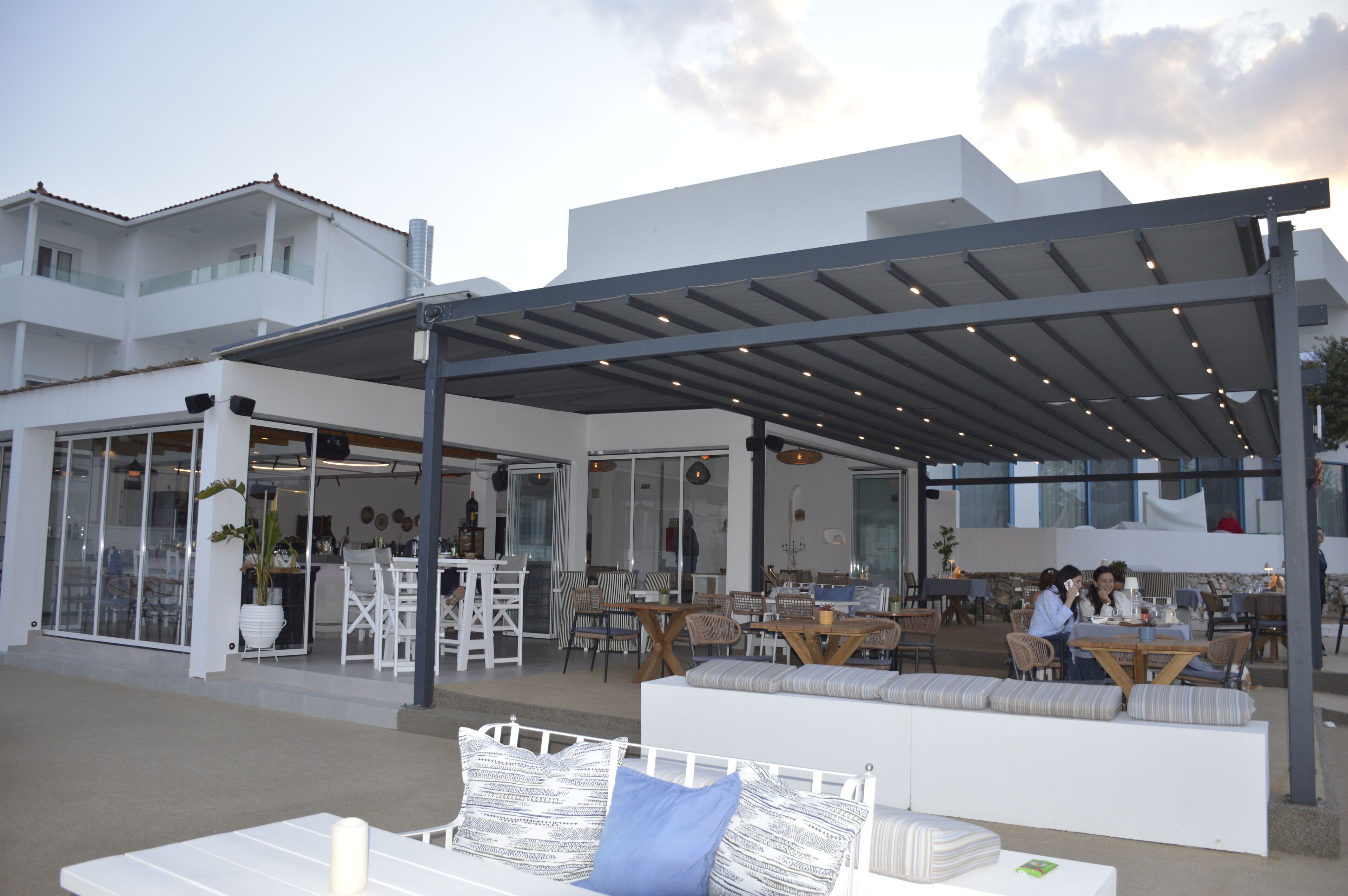 Ein Sitzbereich im Freien mit Tischen und Stühlen, eine überdachte Terrasse und ein weißes Gebäude mit großen Fenstern.
