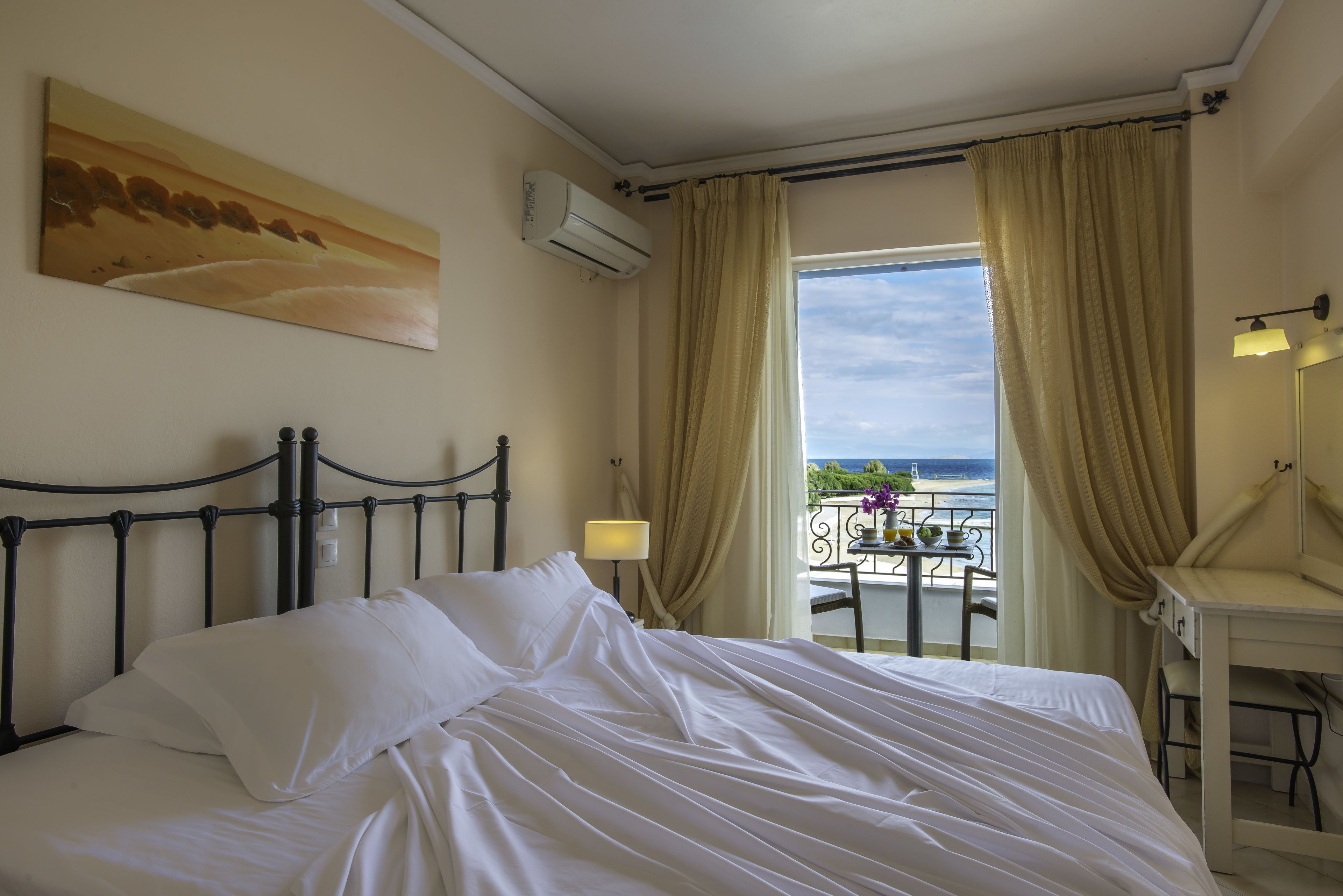 A hotel room with a bed, a painting on the wall, an air conditioner, a balcony with a table and chairs, and a desk with a lamp.