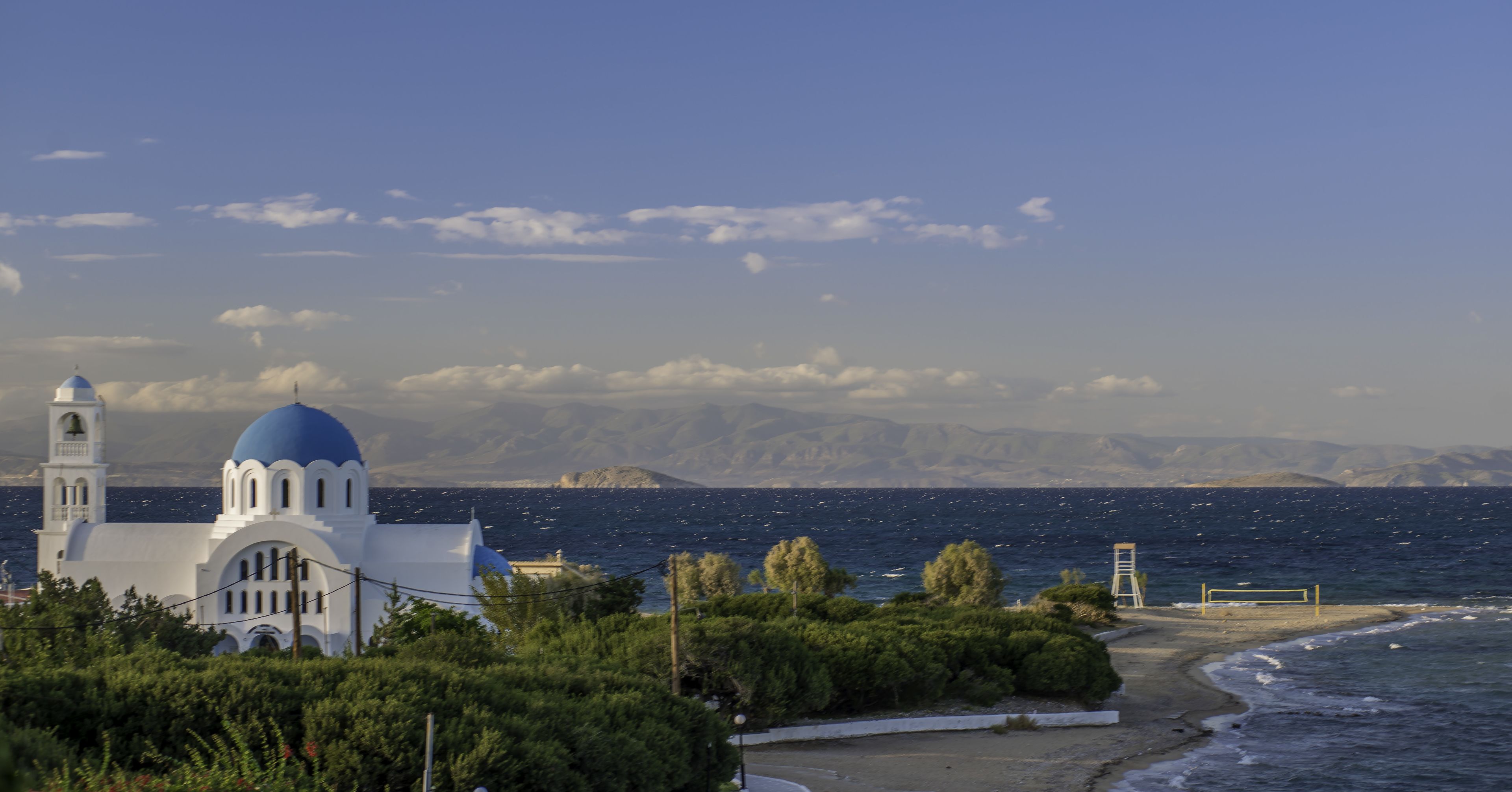 Vista costera con un edificio blanco de cúpulas azules, una playa y montañas al fondo.