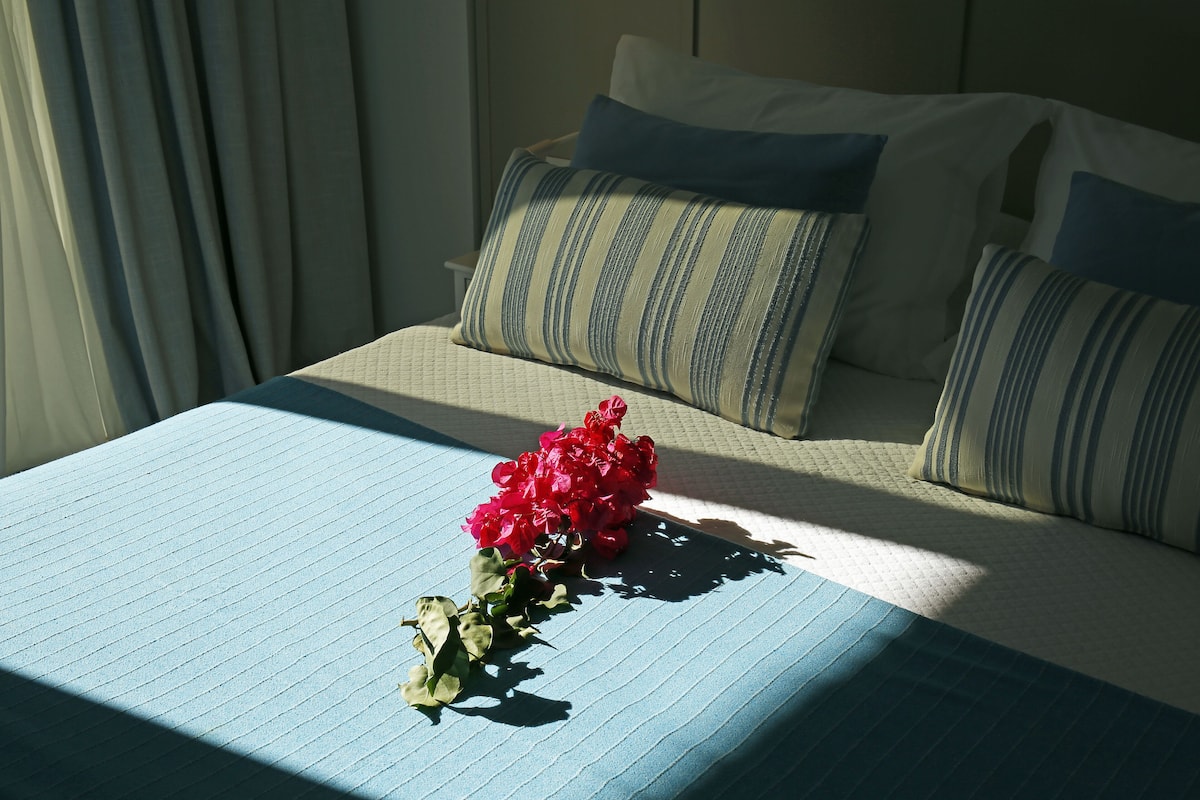 A neatly made bed with a blue bedspread, two pillows with striped patterns, and a bouquet of red flowers on the bed.