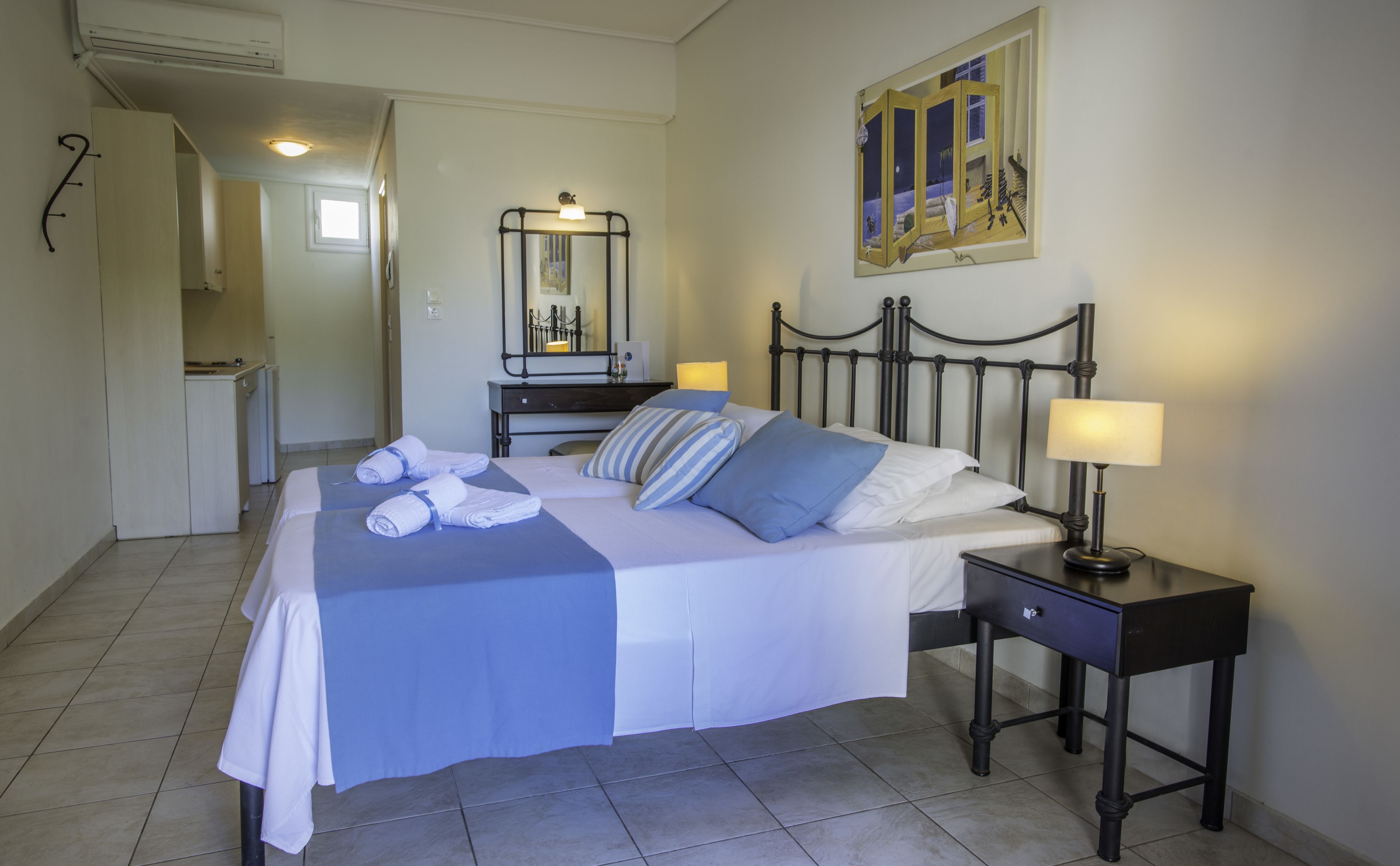 Una cama bien hecha con almohadas y toallas, una mesita de noche con lámpara, un cuadro en la pared y vistas al área de la cocina.
