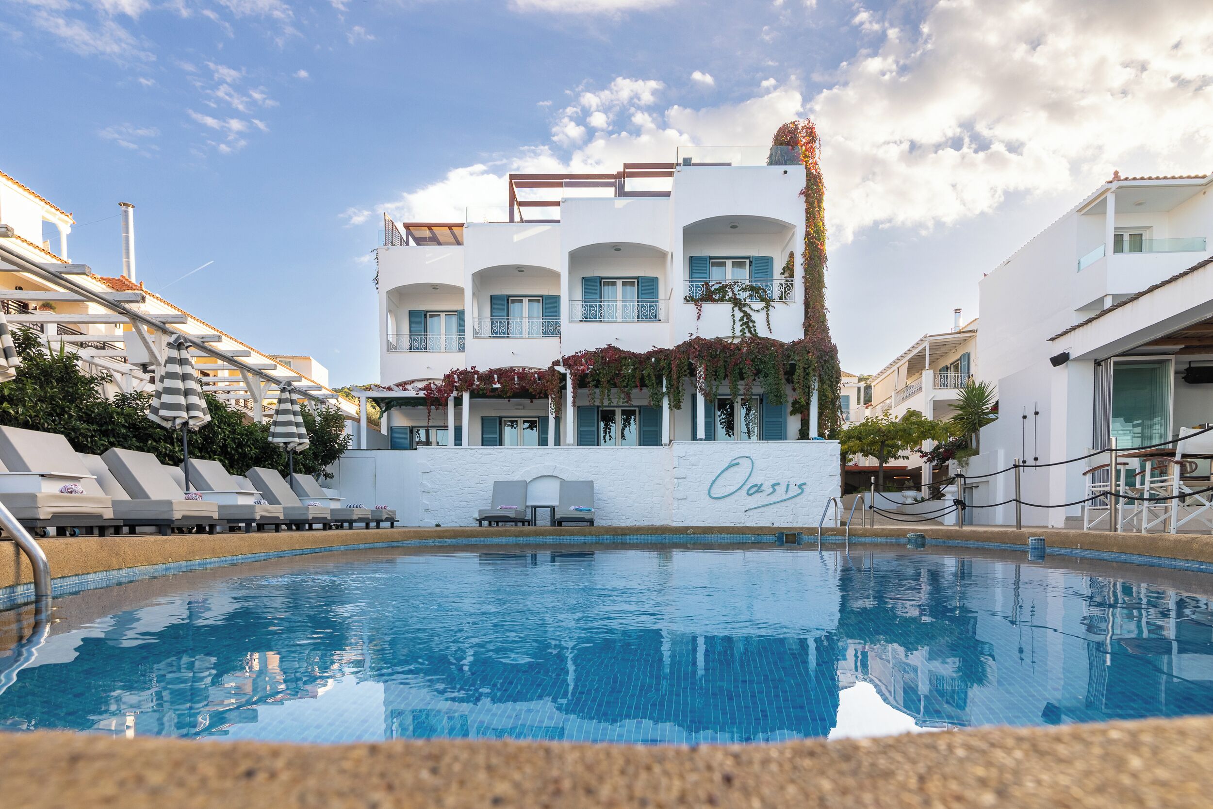 Una piscina con tumbonas y sombrillas, un edificio blanco con balcones y la palabra "Oasis" en la pared.