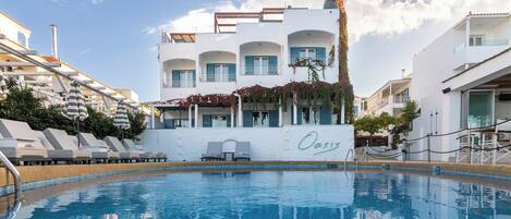 Una piscina con tumbonas y sombrillas, un edificio blanco con balcones y la palabra "Oasis" en la pared.