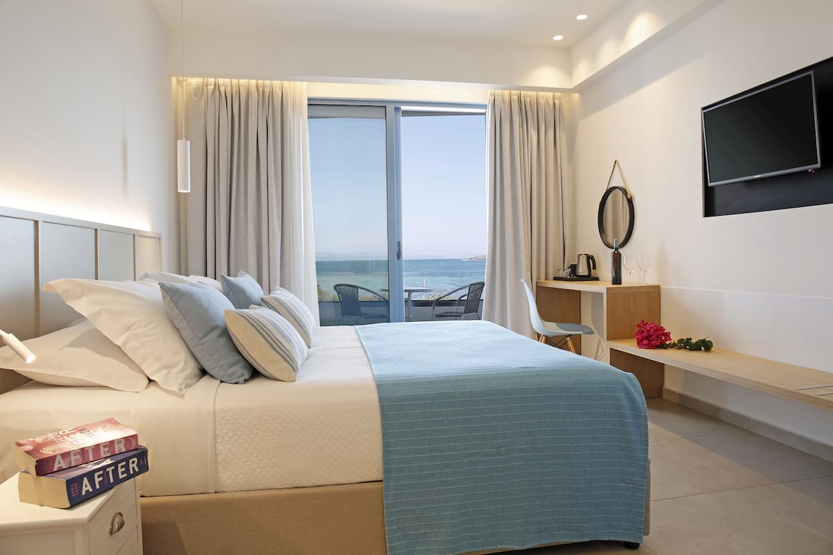 A modern hotel room with a large bed, a desk, and a view of the sea through a sliding door.