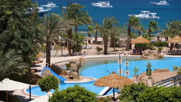 Een resort met een zwembad, palmbomen en boten op het water.