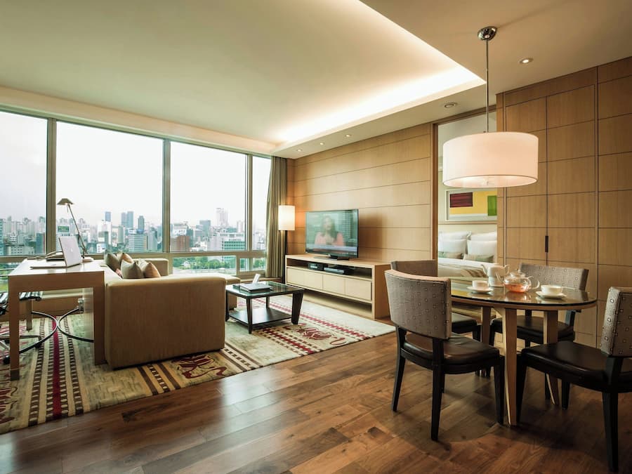 A modern hotel room with a large window offering a city view, a comfortable seating area with a sofa and chairs, a dining table with chairs, and a television on a wooden cabinet.