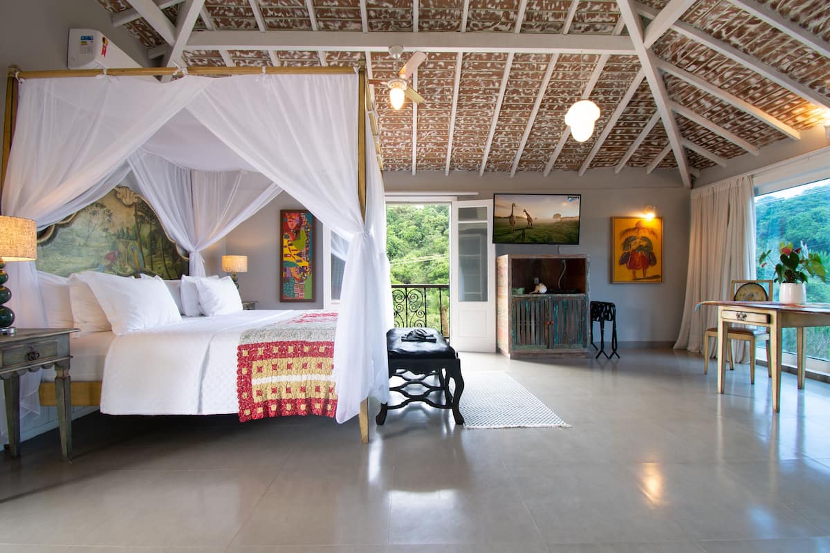 Une chambre spacieuse avec un lit à baldaquin, un coin repas avec une table et des chaises, et un balcon offrant une vue sur la verdure.