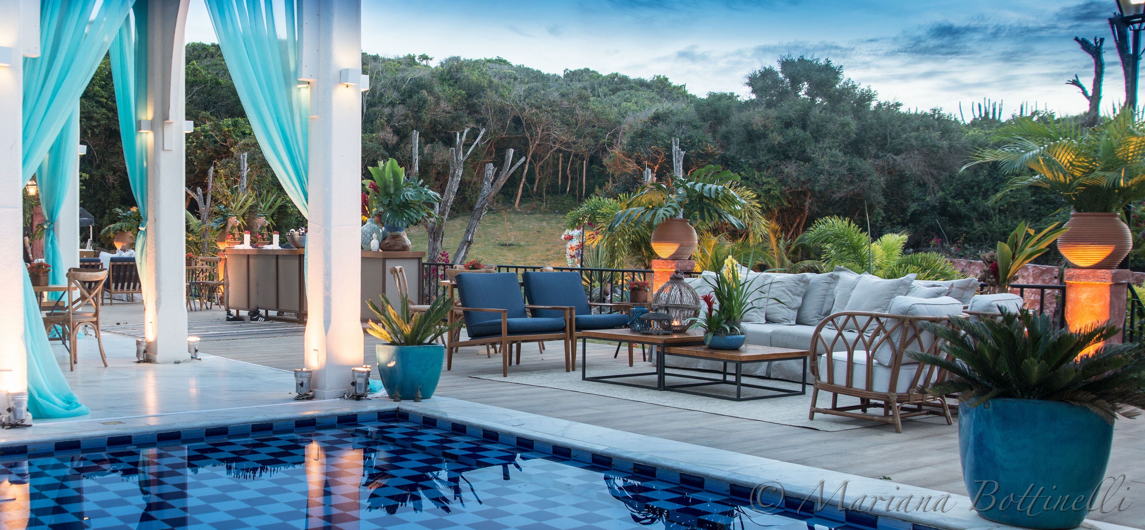 An outdoor pool area with a checkered tile pattern, surrounded by blue and white drapes, wooden furniture, and potted plants.