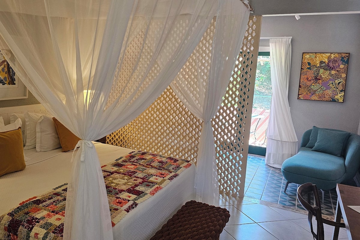 Une chambre à coucher avec un lit, des rideaux blancs, une courtepointe, une chaise et un tableau accroché au mur.