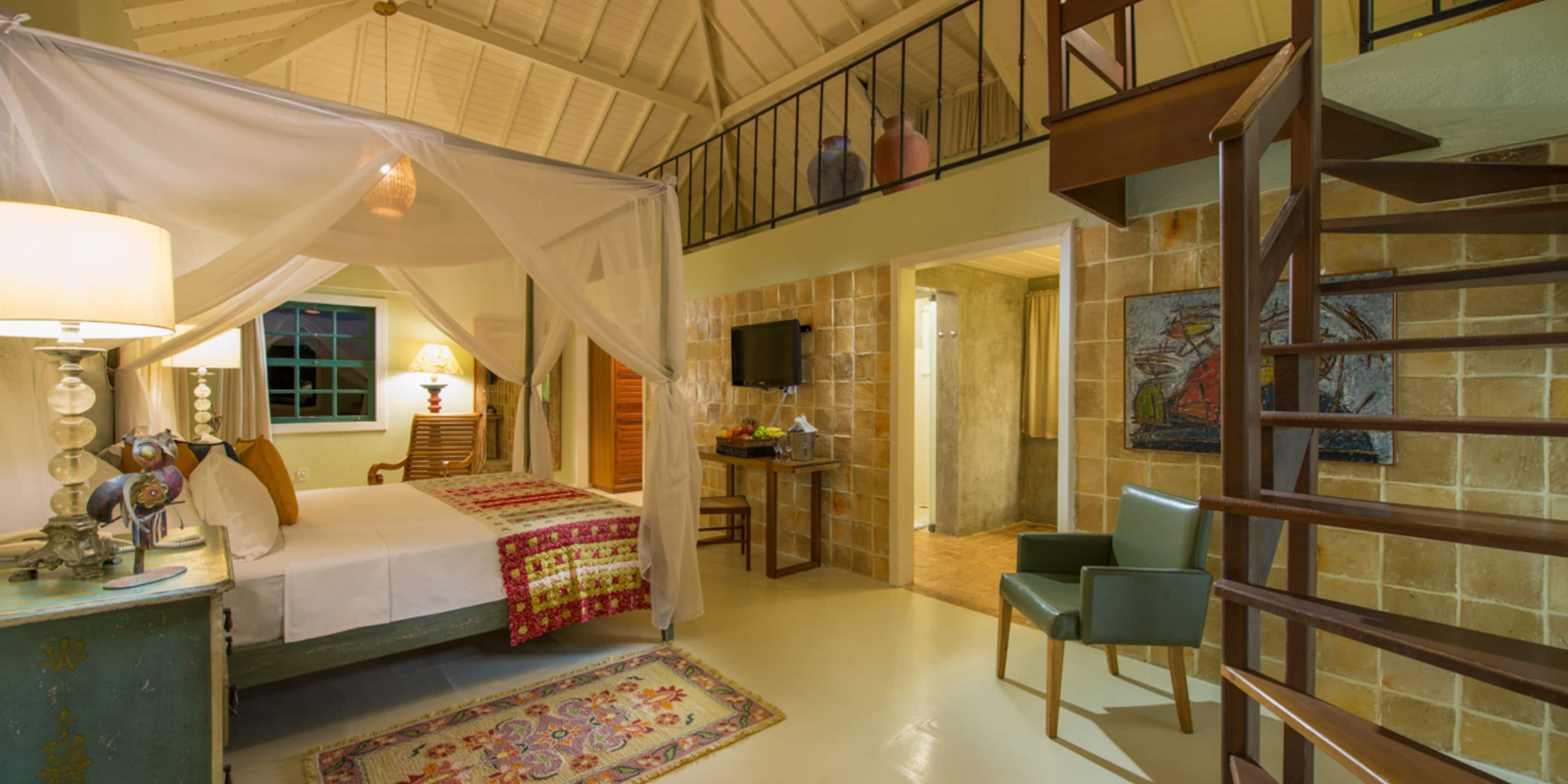 A bedroom with a four-poster bed, a sitting area with a chair, a small table, and a staircase leading to an upper level.
