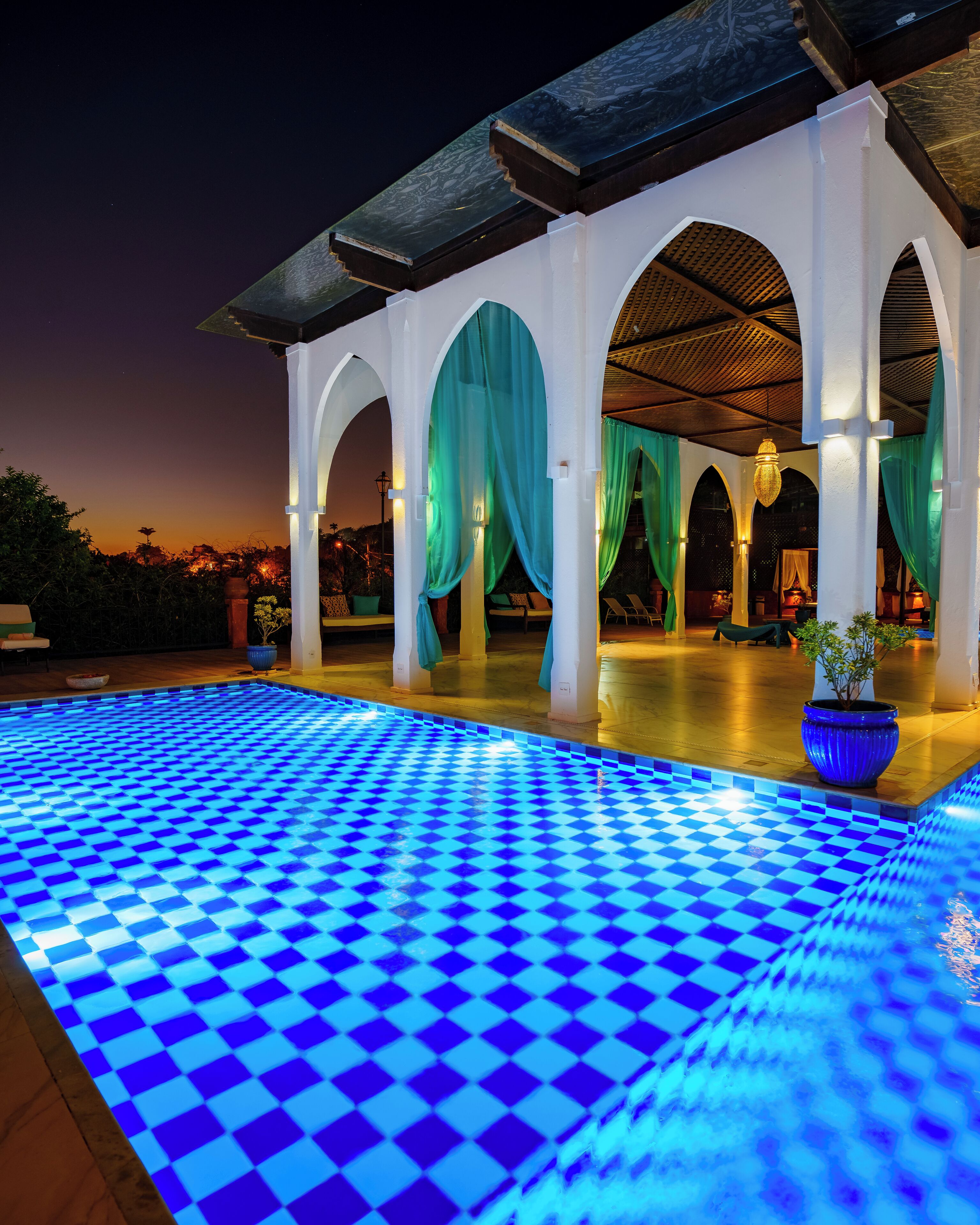 Une piscine aux motifs à carreaux bleus, entourée de piliers blancs et de rideaux verts.