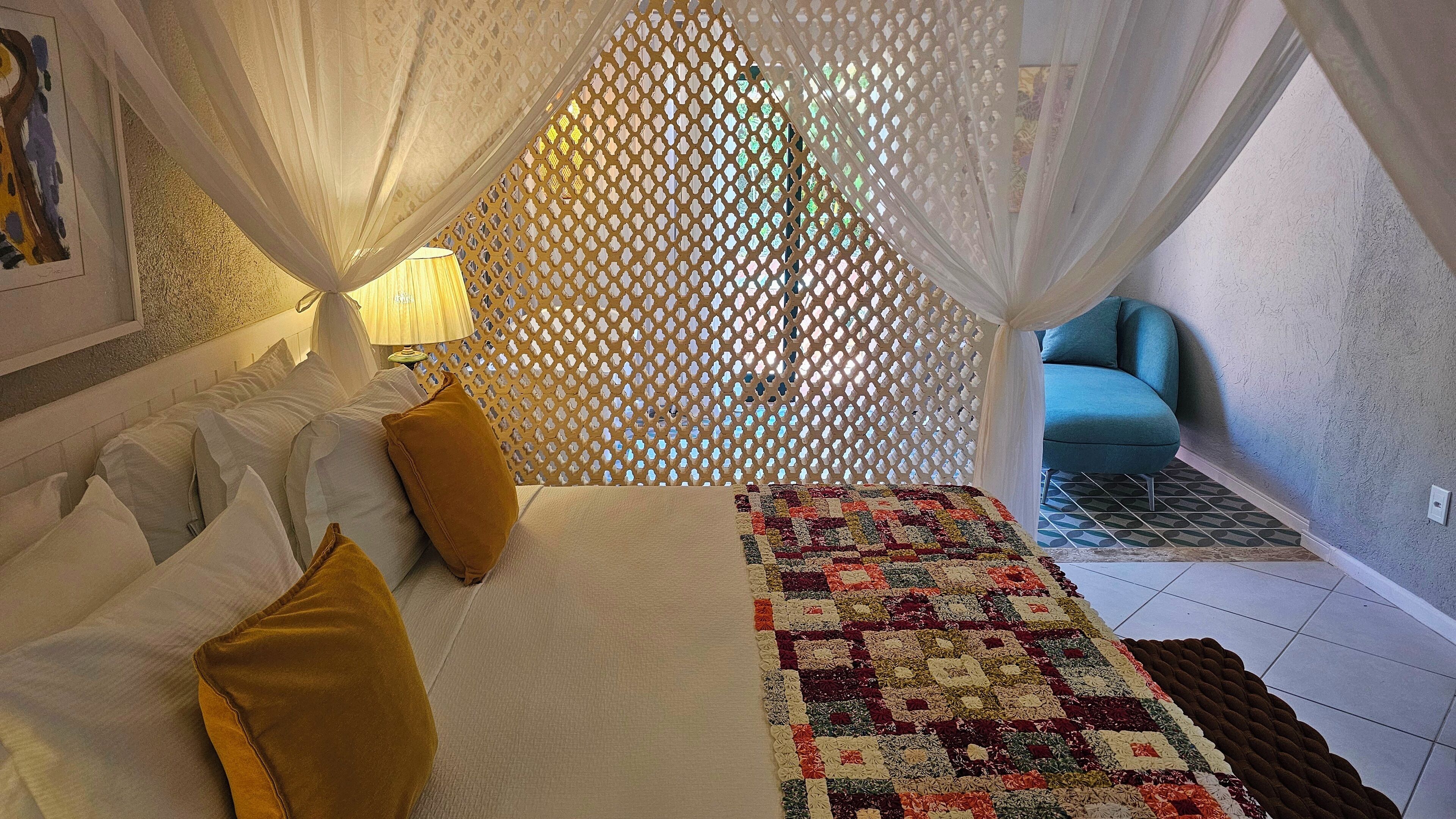 A bedroom with a bed, pillows, a quilt, a nightstand, a lamp, a window with a view, and a blue chair in the corner.
