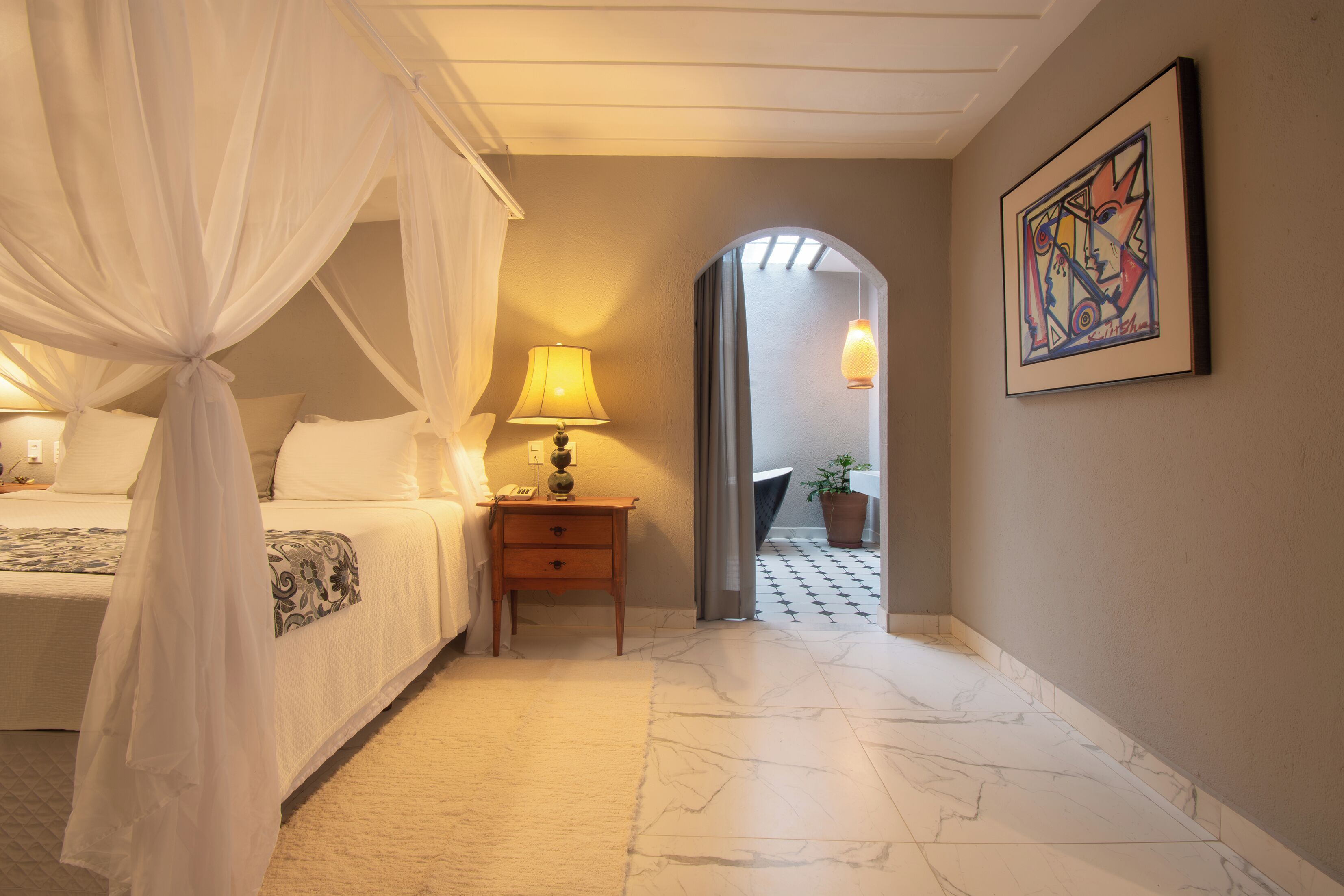 Ein Hotelzimmer mit einem Bett, einem Nachttisch mit Lampe, einem Bild an der Wand und einem Flur, der zu einem anderen Zimmer führt.