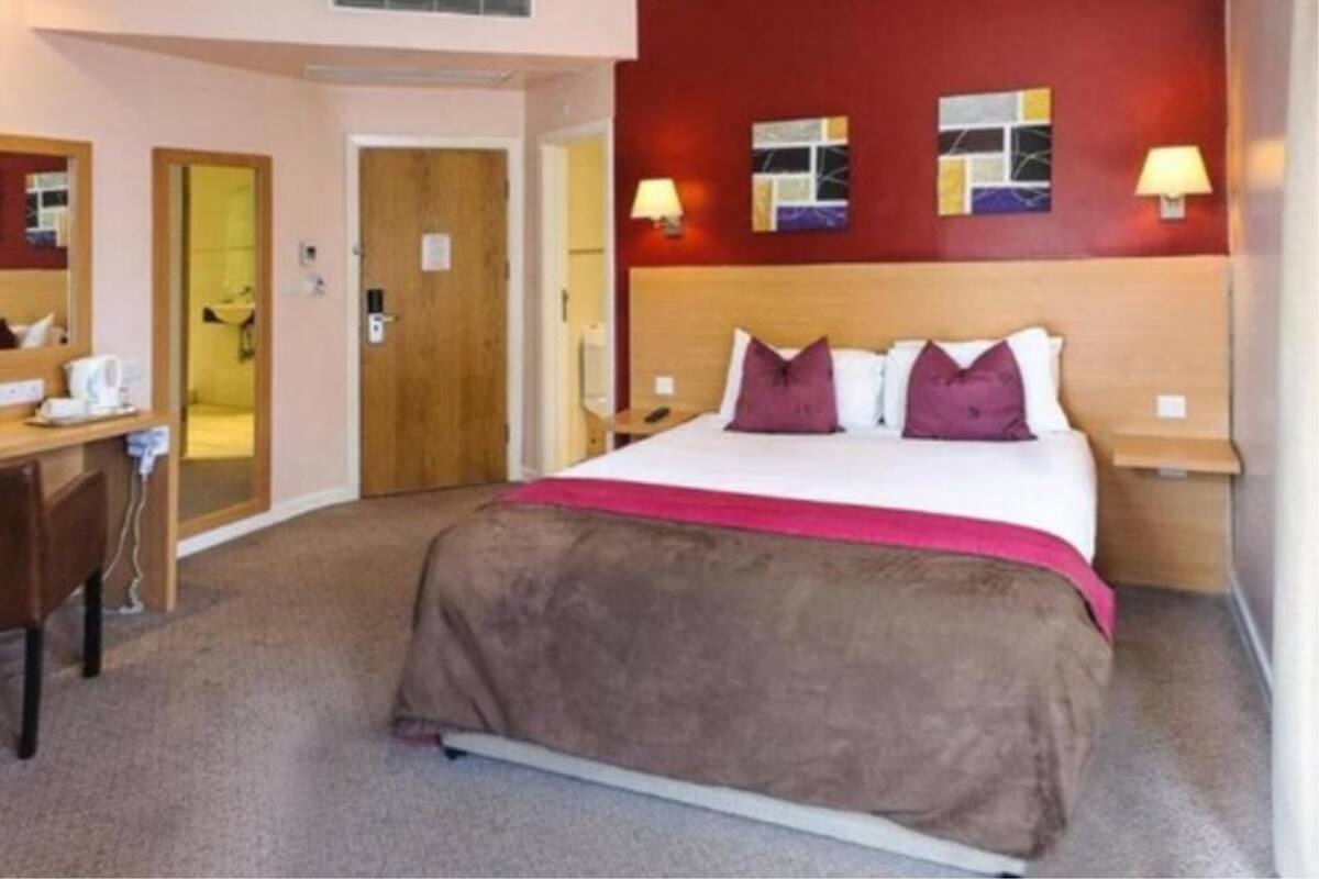 Quarto de hotel com uma cama, uma escrivaninha, uma cadeira, um espelho e uma porta.