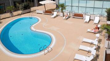A swimming pool with a wavy edge, surrounded by lounge chairs and umbrellas, with a building in the background.
