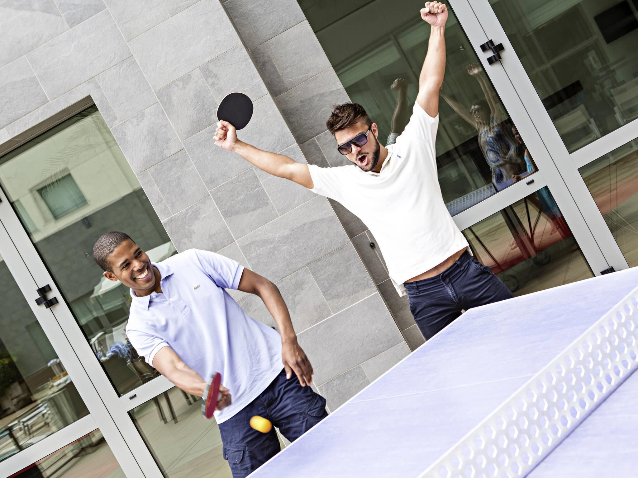 Twee mensen spelen buiten tafeltennis, met een modern gebouw op de achtergrond.