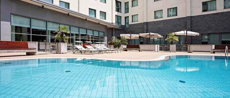 A swimming pool with lounge chairs and umbrellas, surrounded by a building with multiple floors and large windows.