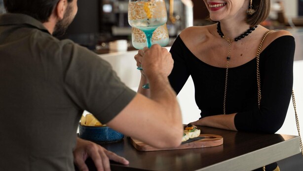 Una persona sosteniendo un vaso con una bebida azul, sentada en una mesa con un plato de comida y un teléfono inteligente.