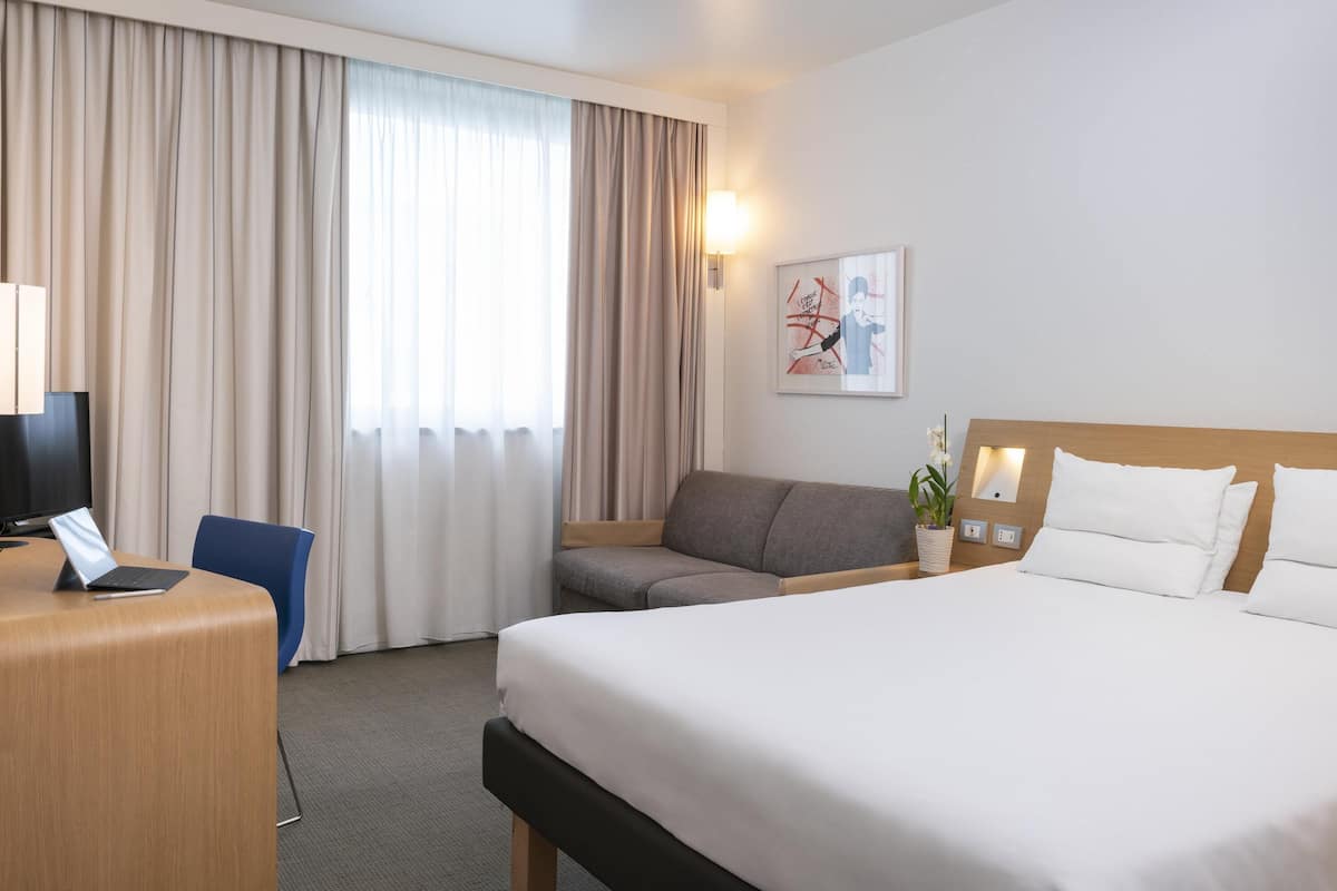 A hotel room with a bed, a desk with a laptop, a chair, a sofa, a window with curtains, and a wall-mounted lamp.