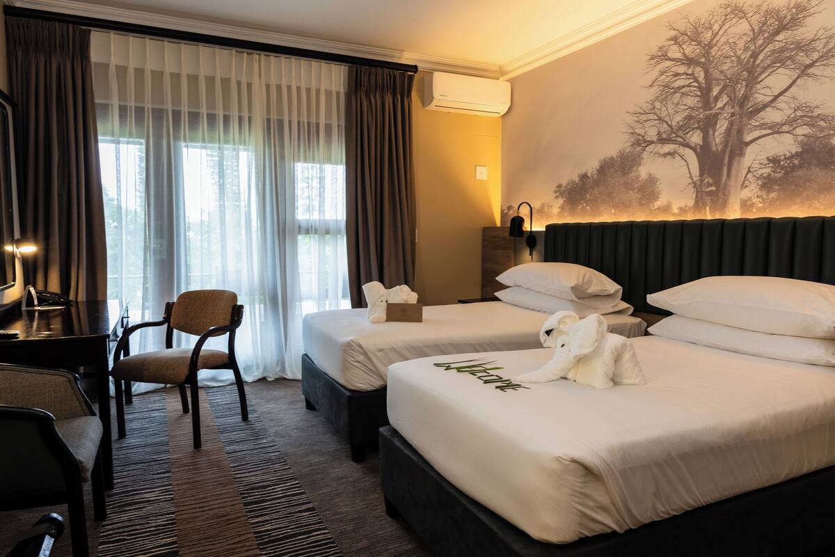 Ein Hotelzimmer mit zwei Betten, einem Schreibtisch, einem Stuhl und einem großen Fenster mit Vorhängen.