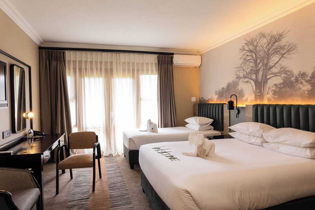 Ein Hotelzimmer mit zwei Betten, einem Schreibtisch, einem Stuhl und einem großen Fenster mit Vorhängen.