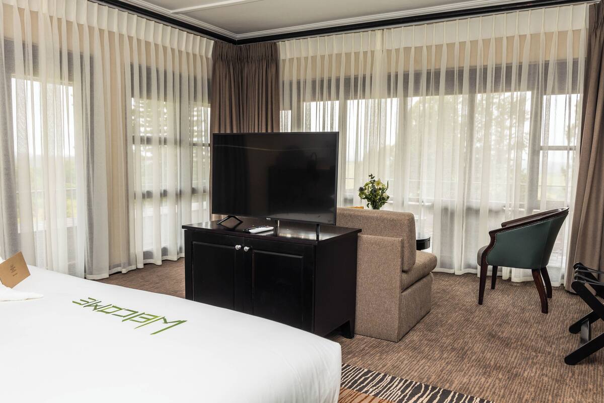 Ein Hotelzimmer mit einem Bett, einem Fernseher auf einem schwarzen Schrank, einem Sofa, einem Sessel und durchscheinenden Vorhängen.