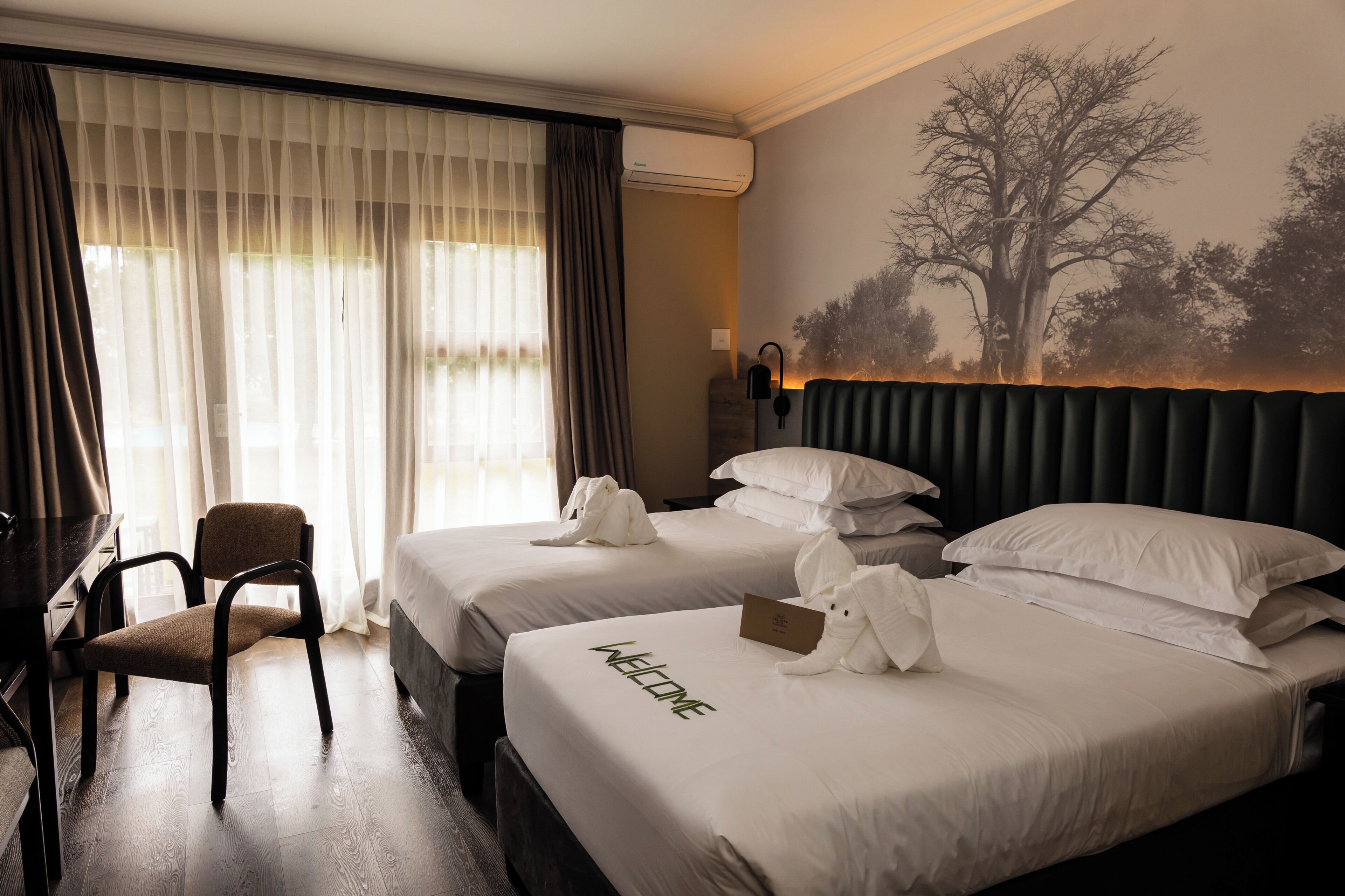 Ein Hotelzimmer mit zwei Betten, einem Schreibtisch, einem Stuhl und einem großen Fenster mit Vorhängen.
