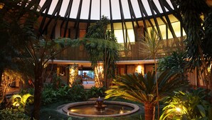 A spacious indoor garden with a central fountain, surrounded by various tropical plants and a high ceiling with wooden beams.