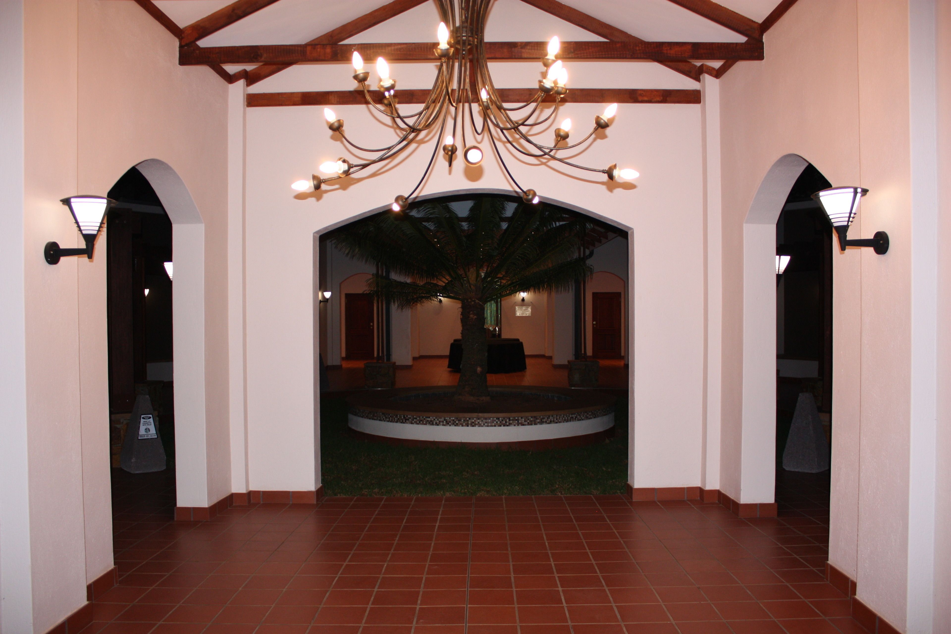 A lobby with a large chandelier, a palm tree, and two archways with wall sconces.