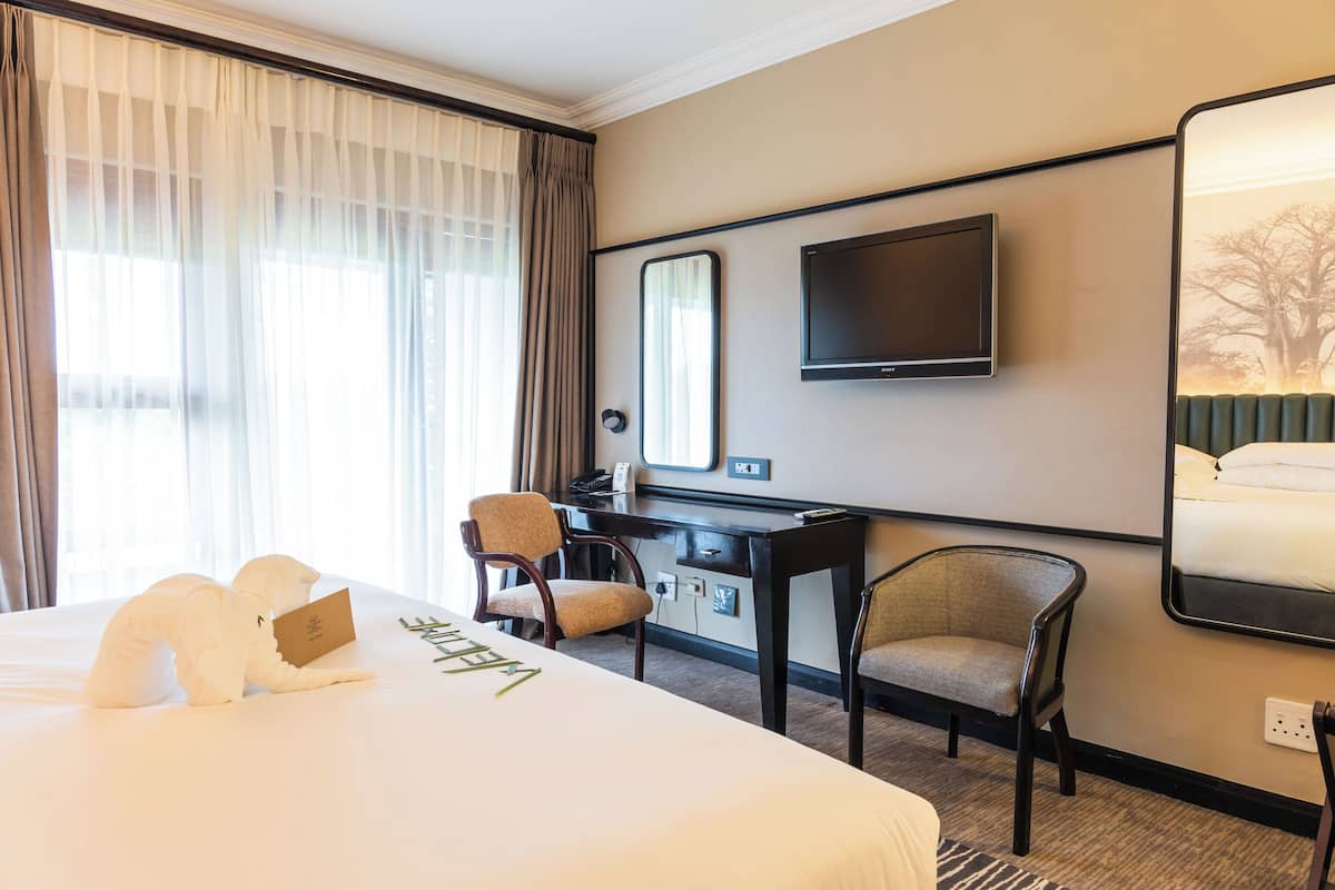 Ein Hotelzimmer mit einem großen Bett, einem Schreibtisch, zwei Stühlen, einem Flachbildfernseher und einem großen Fenster mit Vorhängen.