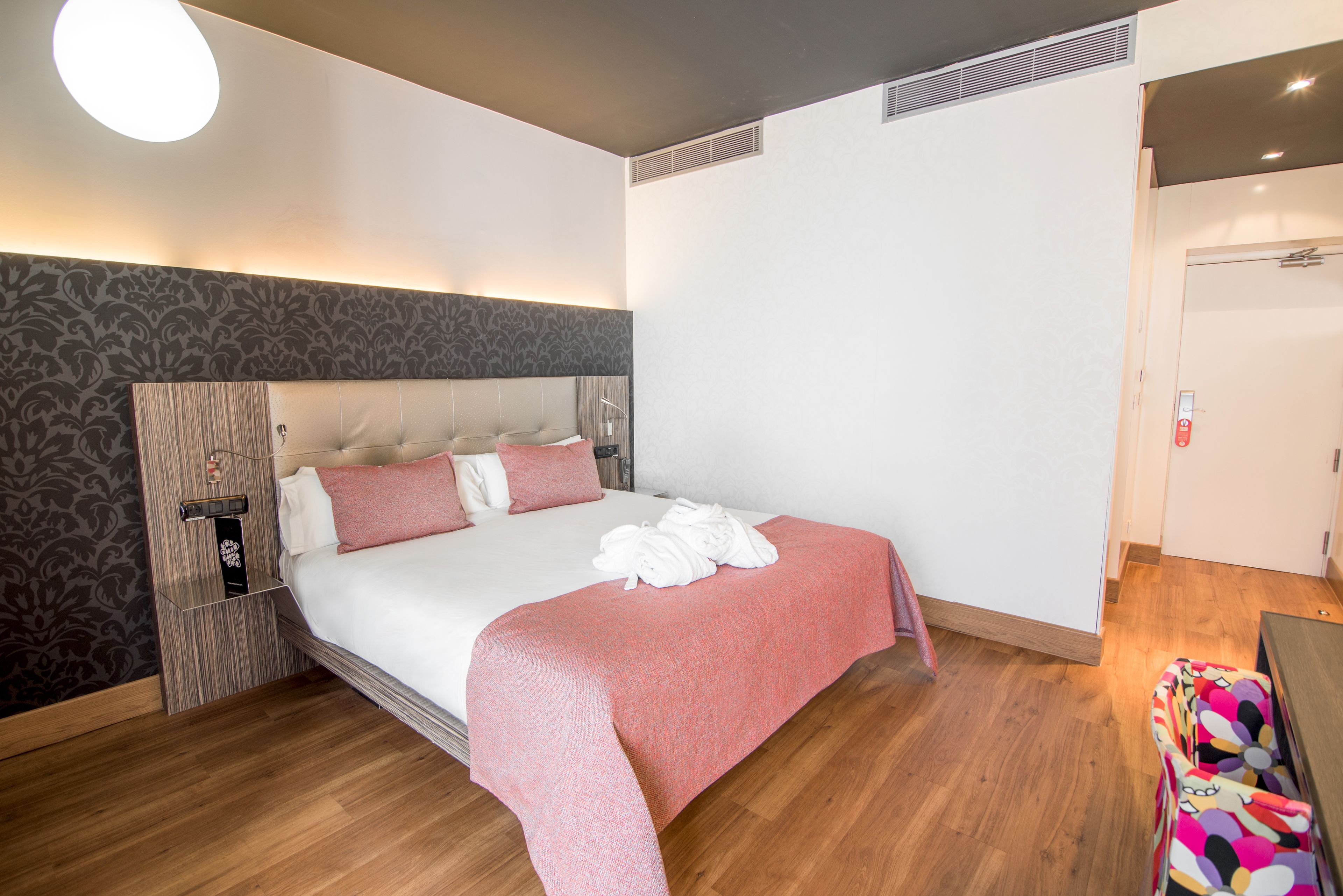 A hotel room with a large bed, a wooden headboard, a bedside table, and a wall-mounted heater.