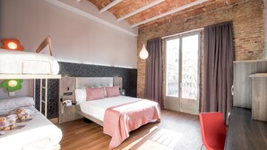 A modern hotel room with a large bed, a desk, and a red chair. There is a window with curtains and a view of the city. - Petit Palace Museum (Barcelona)