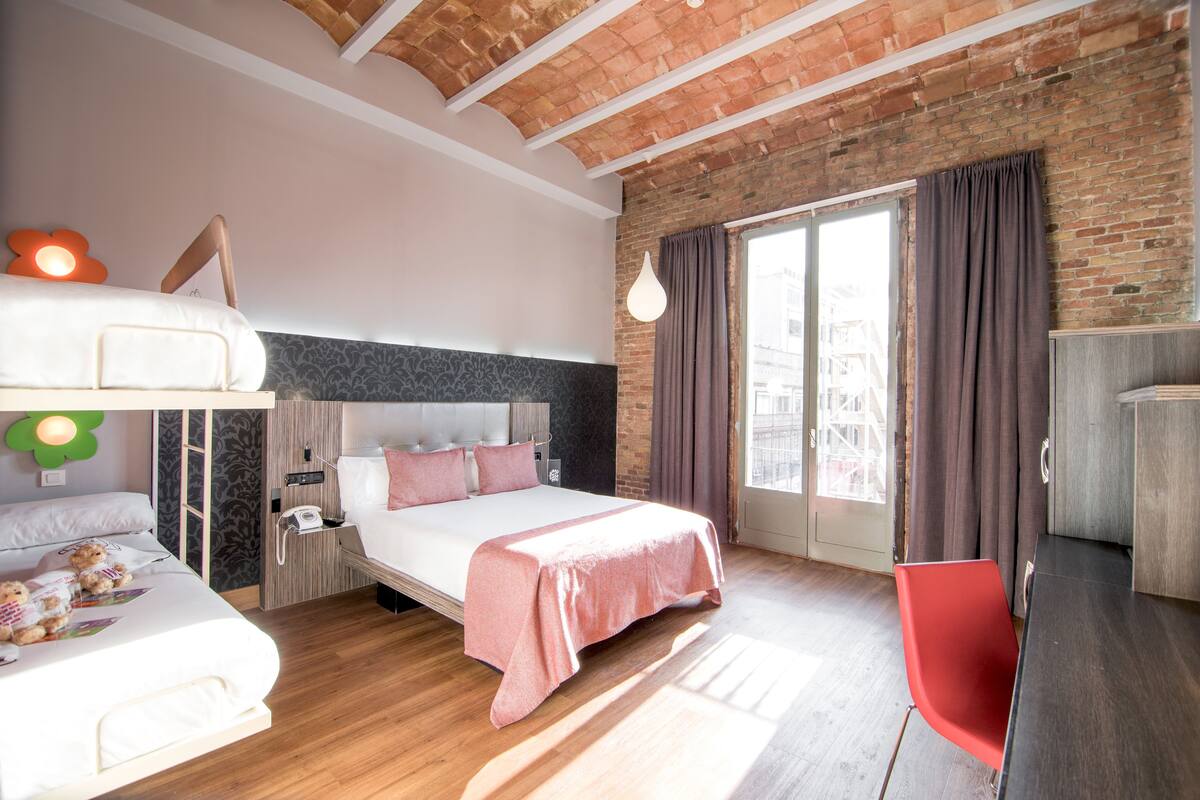 A modern hotel room with a brick wall, a large bed with a pink bedspread, a red chair, and a desk.