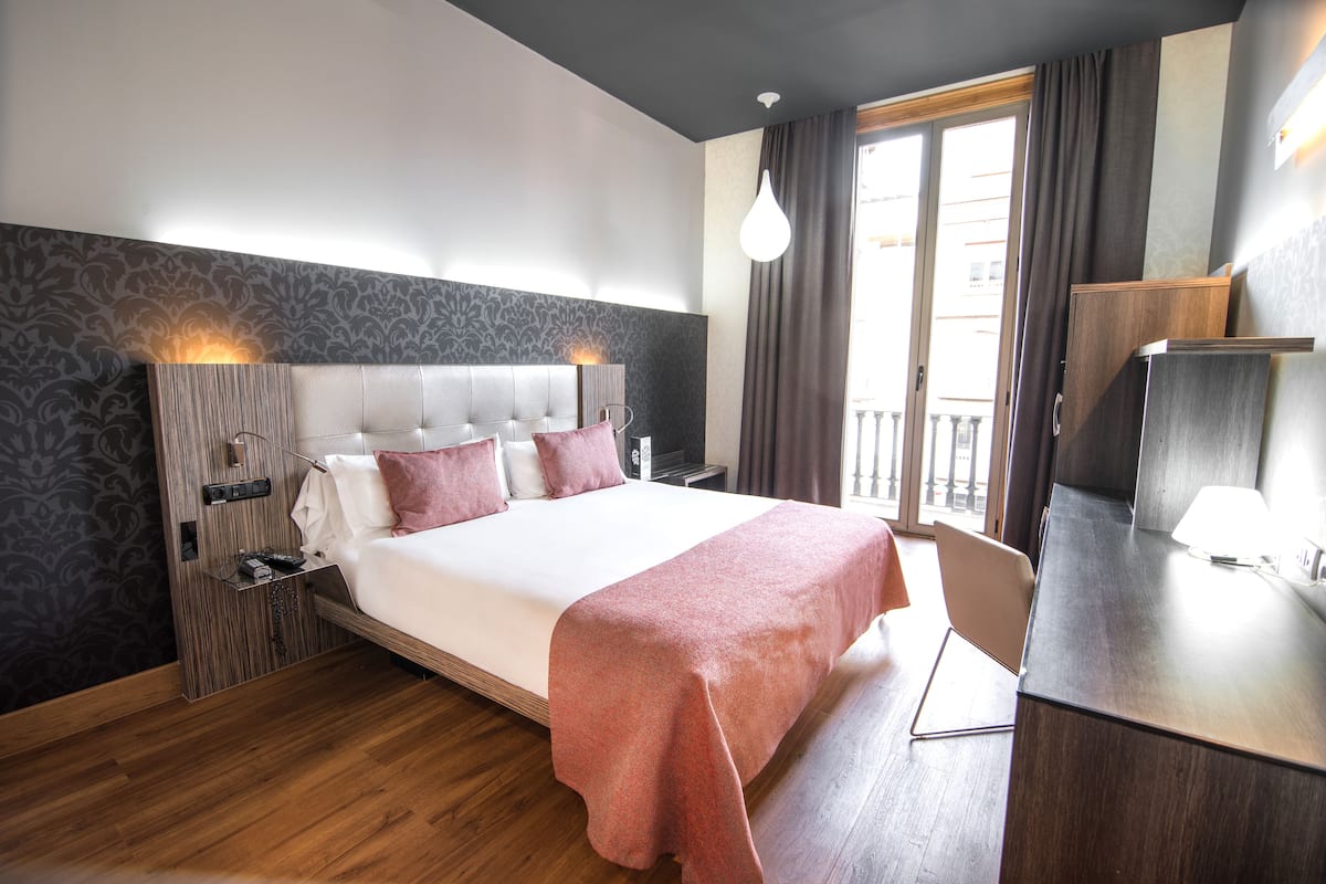 A modern hotel room with a large bed, wooden flooring, a desk, and a balcony with curtains.
